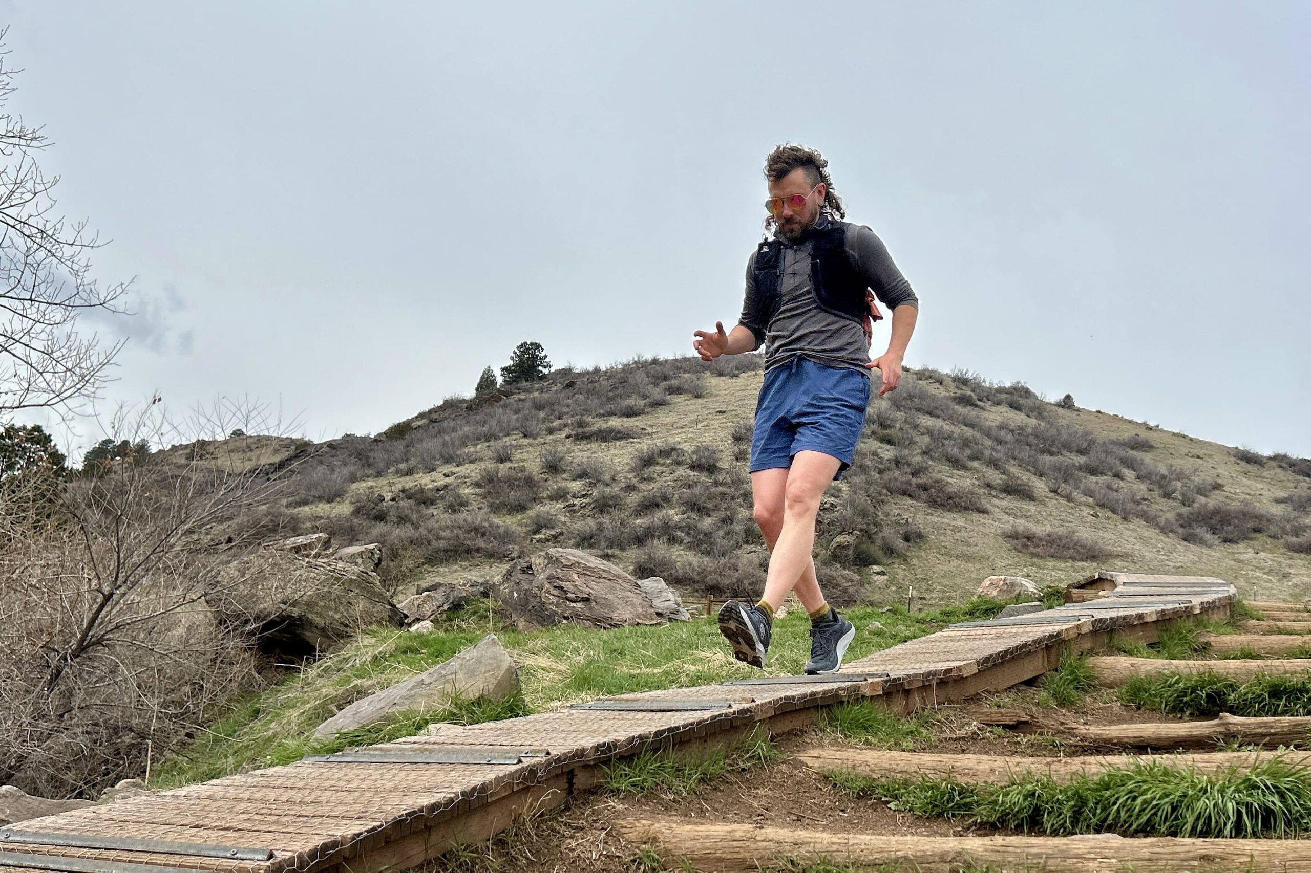 A man runs down a wooden board on a trail in a hilly wilderness setting.