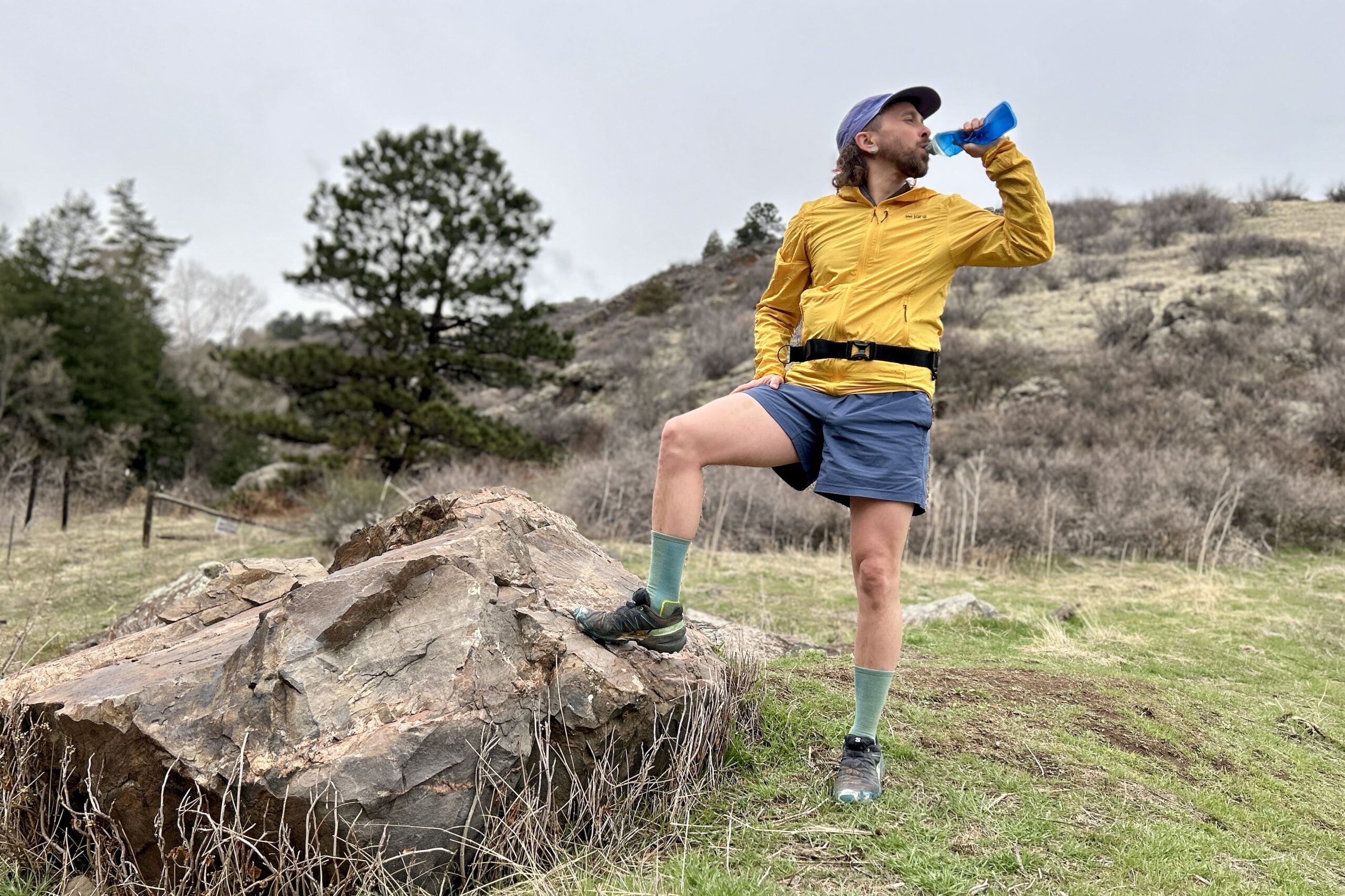 A man stands in a mountain setting drinking water out of a bottle with one leg up on a rock.
