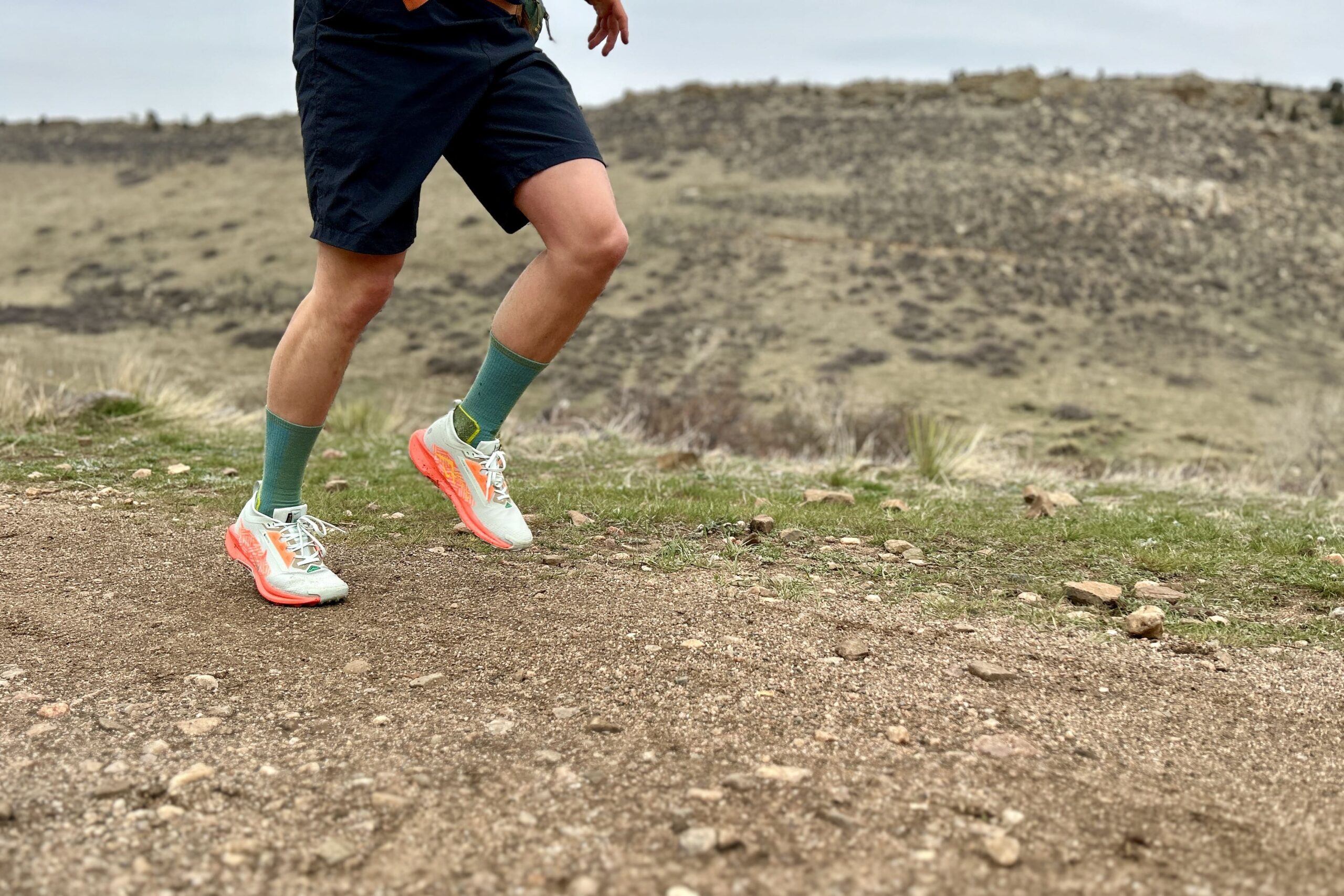 A person from the waist down runs on a dirt road in a mountain setting.