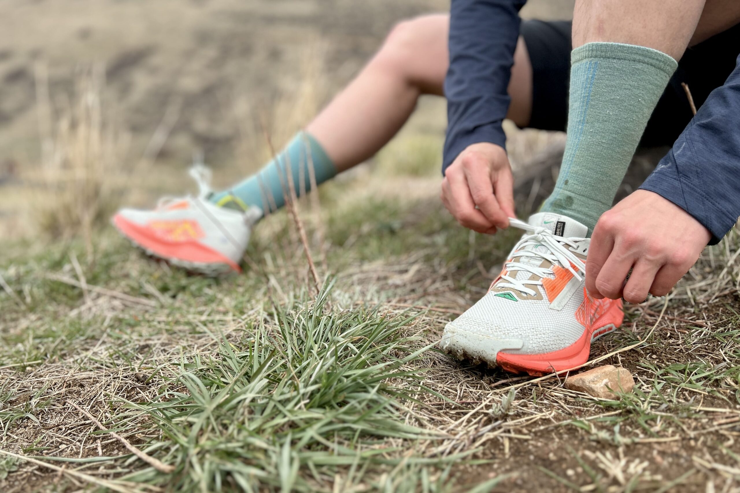 Close up from the waist down of a person tying a pair of shoes as they sit on the ground in a grassy patch.