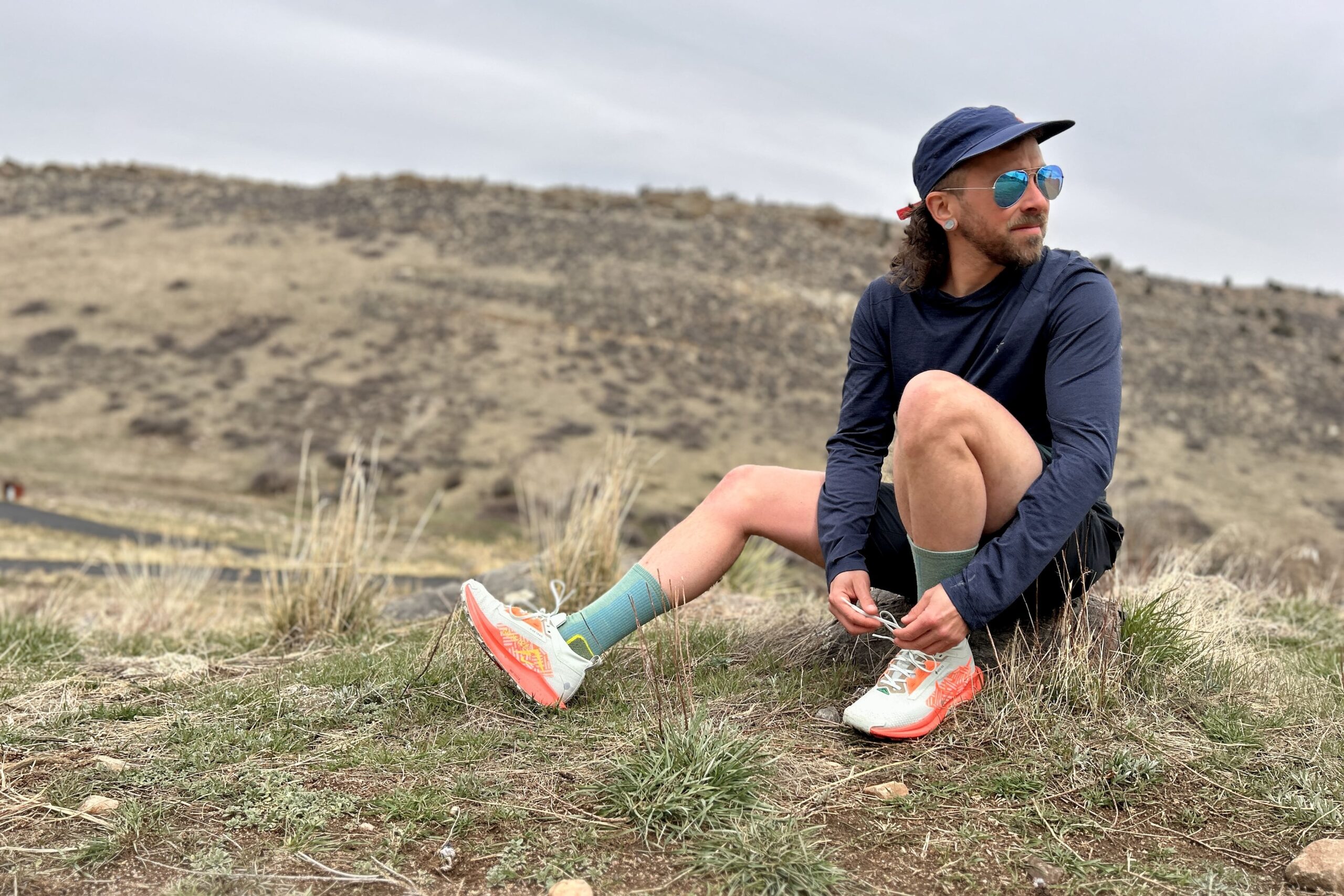 A man in trail running attire and blue sunglasses ties his shoes and looks off in the distance.