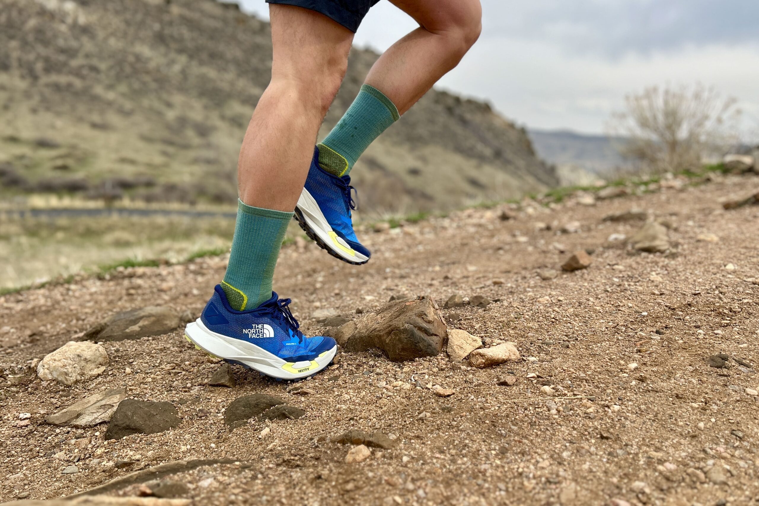 Close up image from teh waist down of a man running uphill in shorts and trail runners.