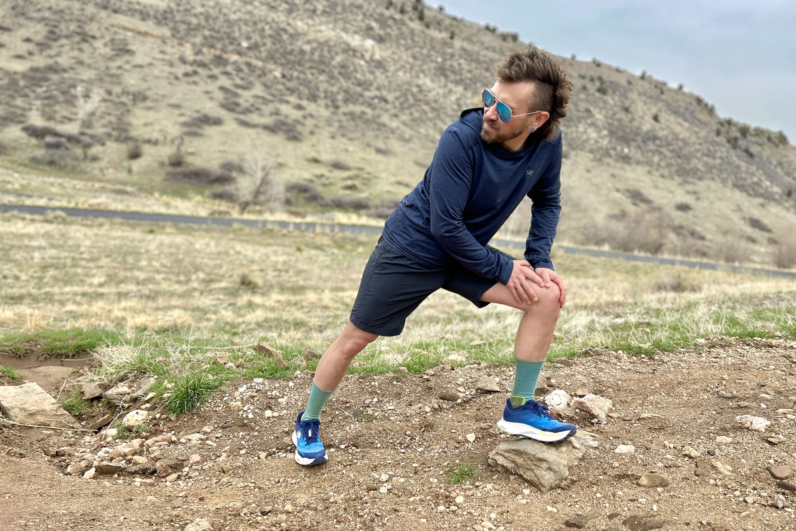 A man leans over and stretches in a mountain setting, looking off in the distance.