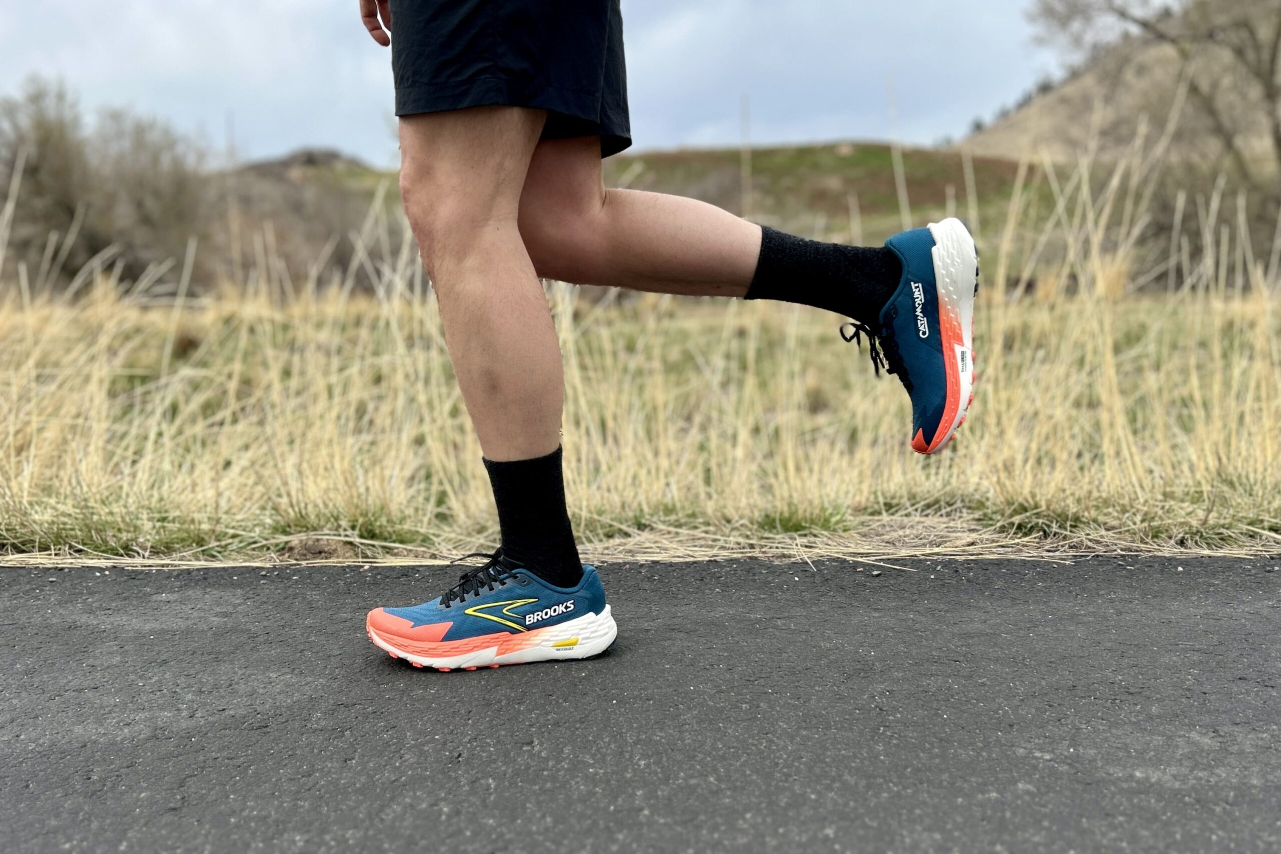 Close up image from the waist down of a person in trail running shoes and shorts running on a road with tall grass at the edge.