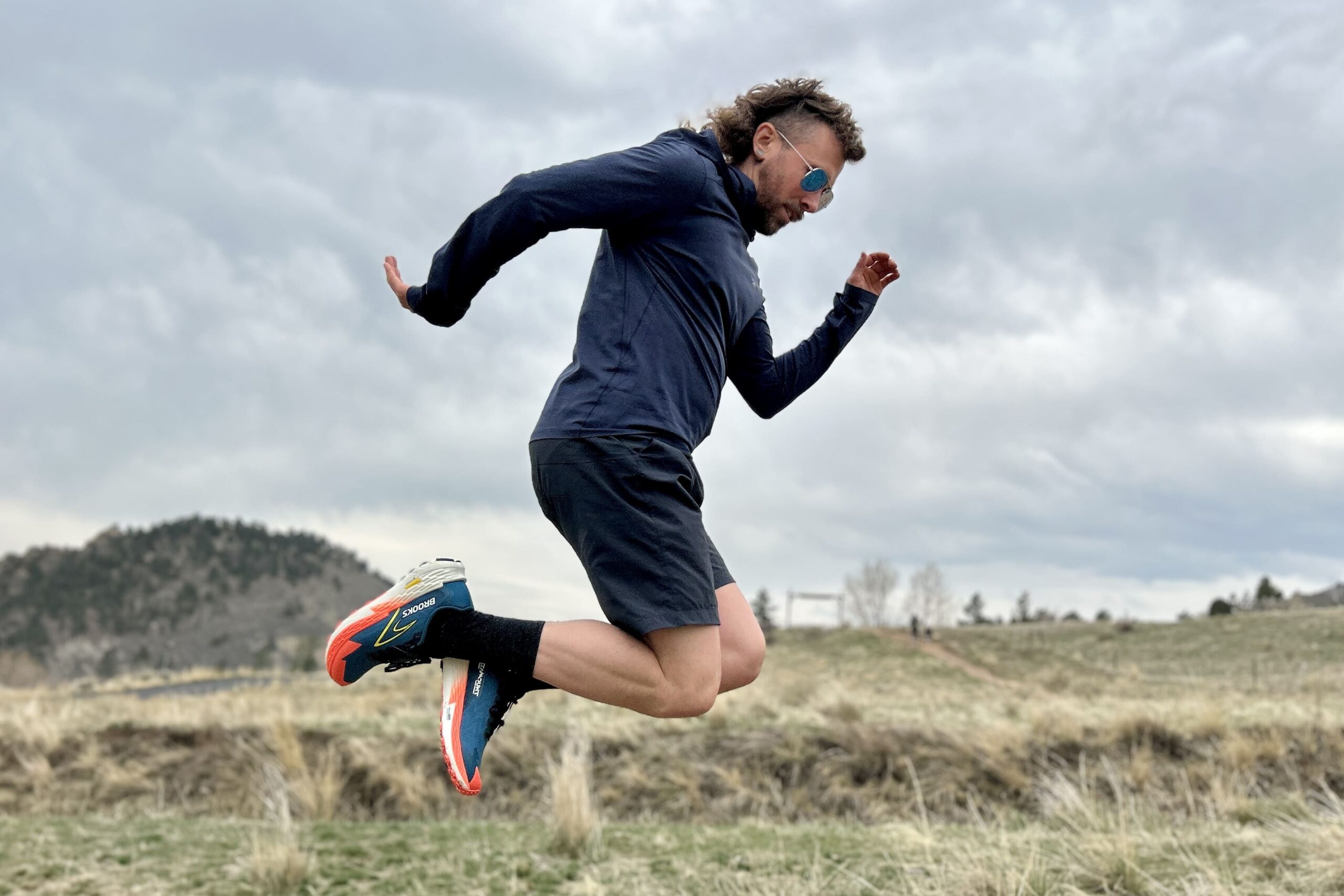 A man jumping in the air wearing trail running attire under a moody, cloudy sky.