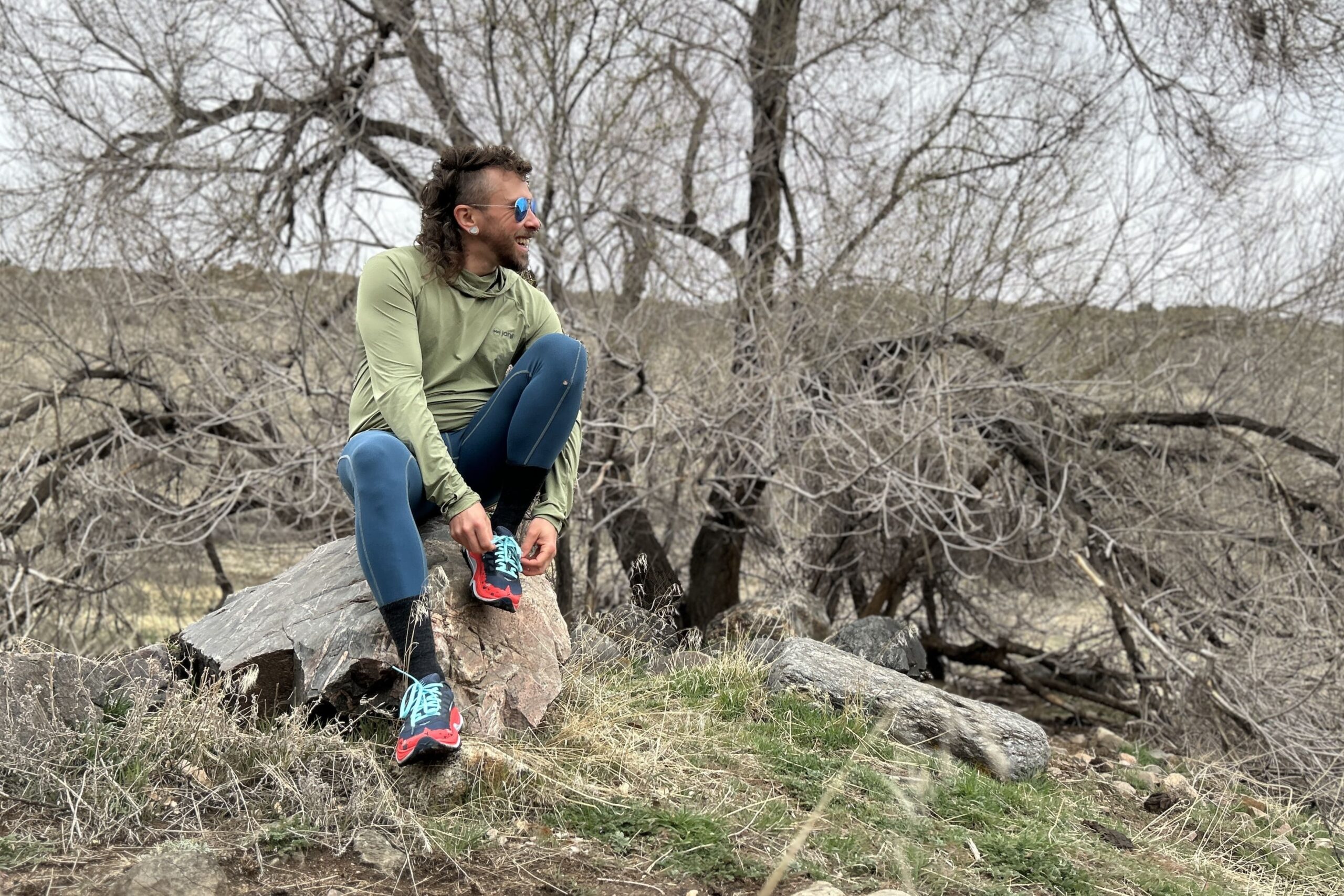 A man sits on a rock in a wooded landscape tying his shoes and looking off in the distance.