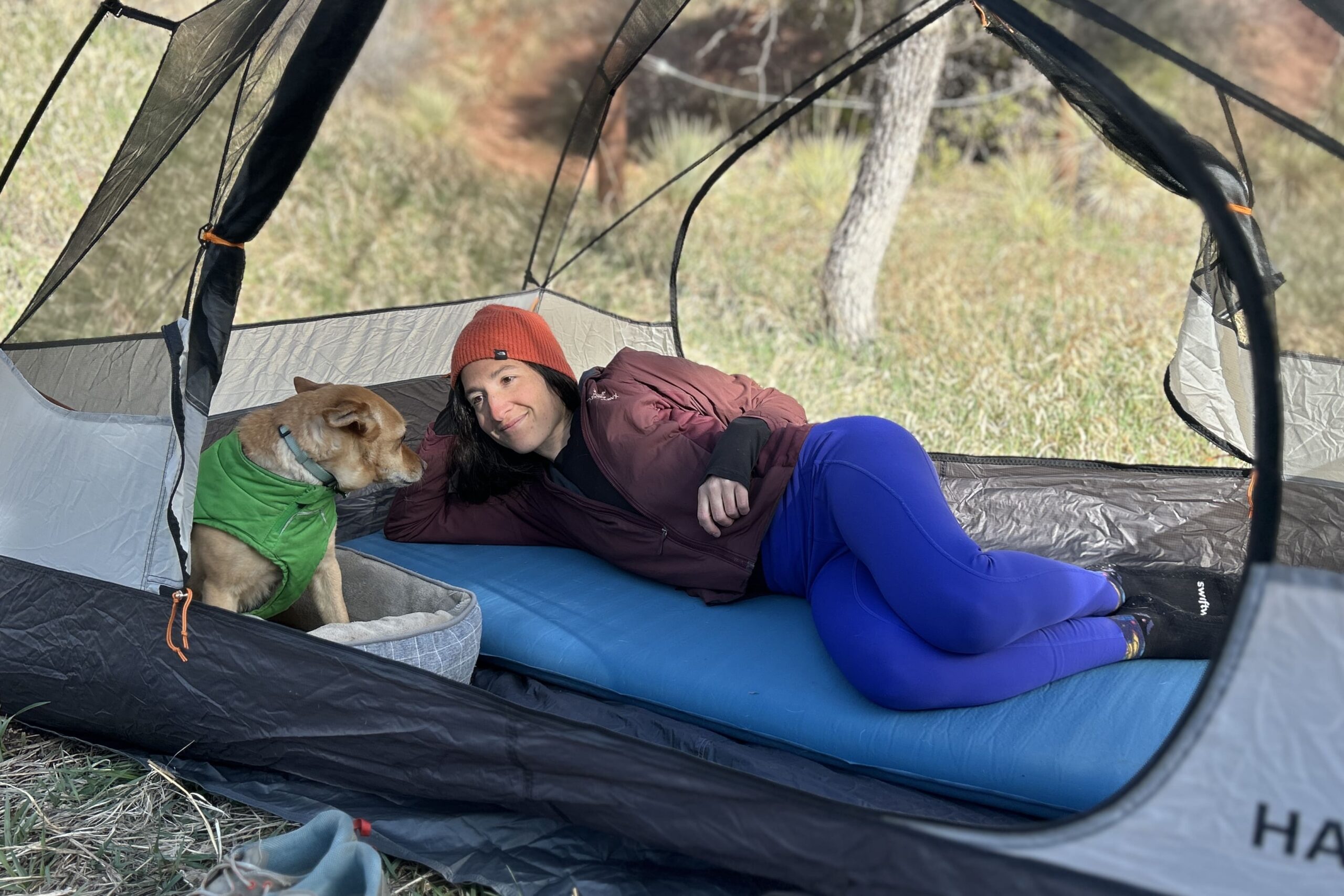 A woman lays on her side on a camping mattress in a tent smiling at her dog, who is laying in a dog bed.
