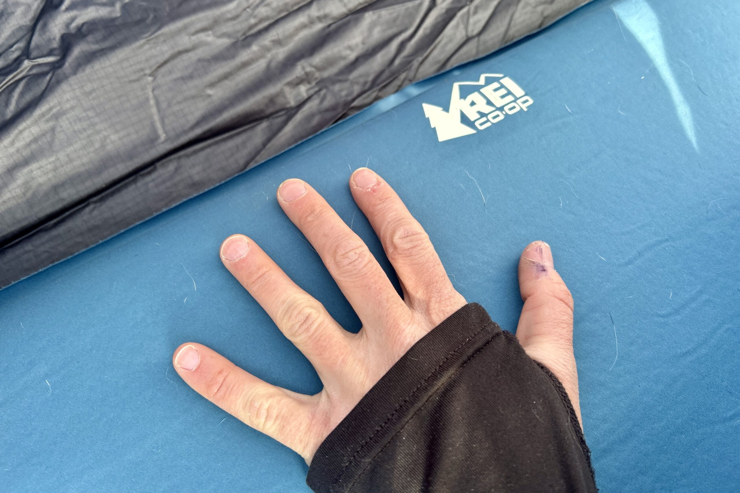 A person's hand rests on a camping mattress with the REI logo visible in the corner.