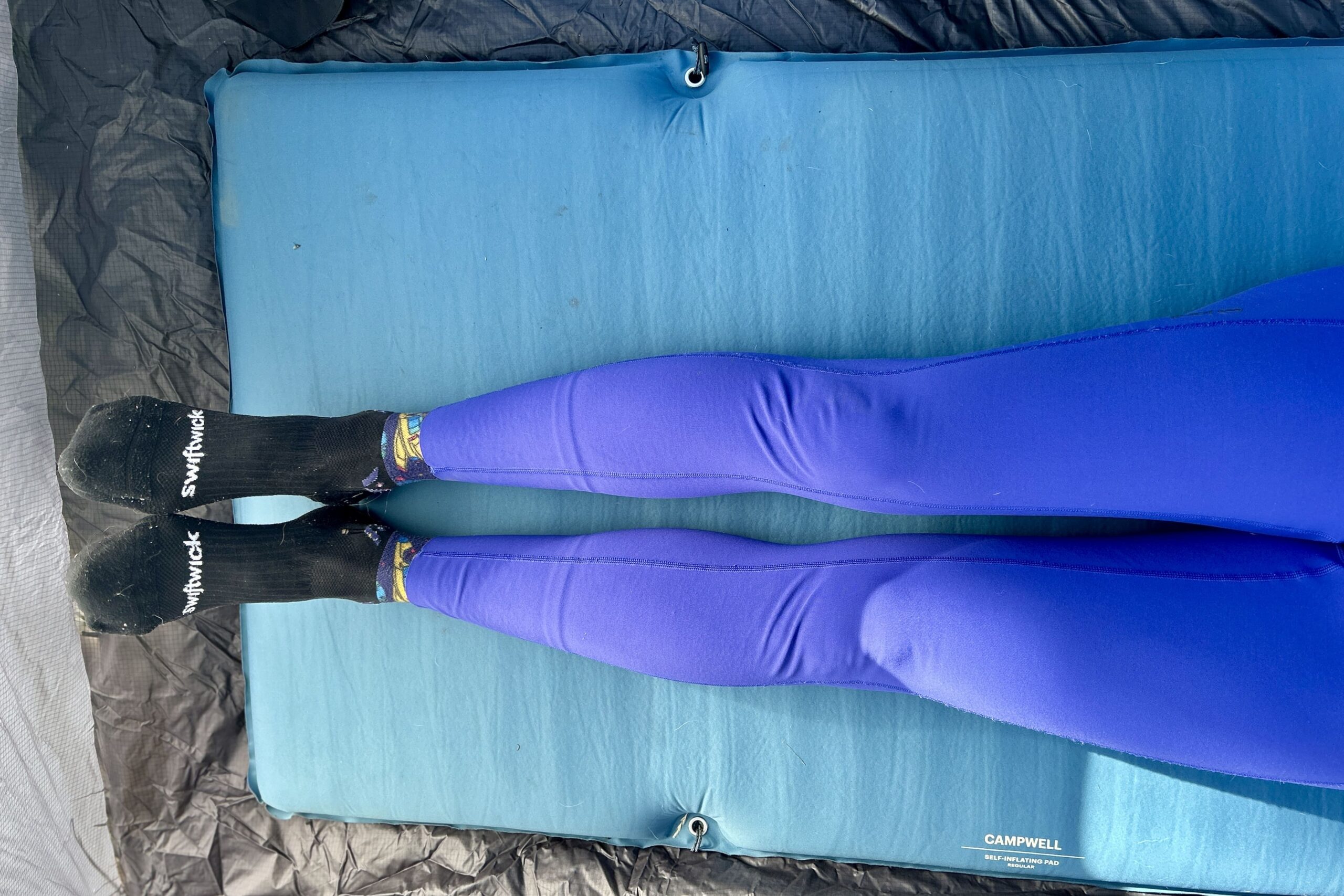 Top-down view of a person's legs wearing leggings and socks and resting on a camping mattress in a tent.