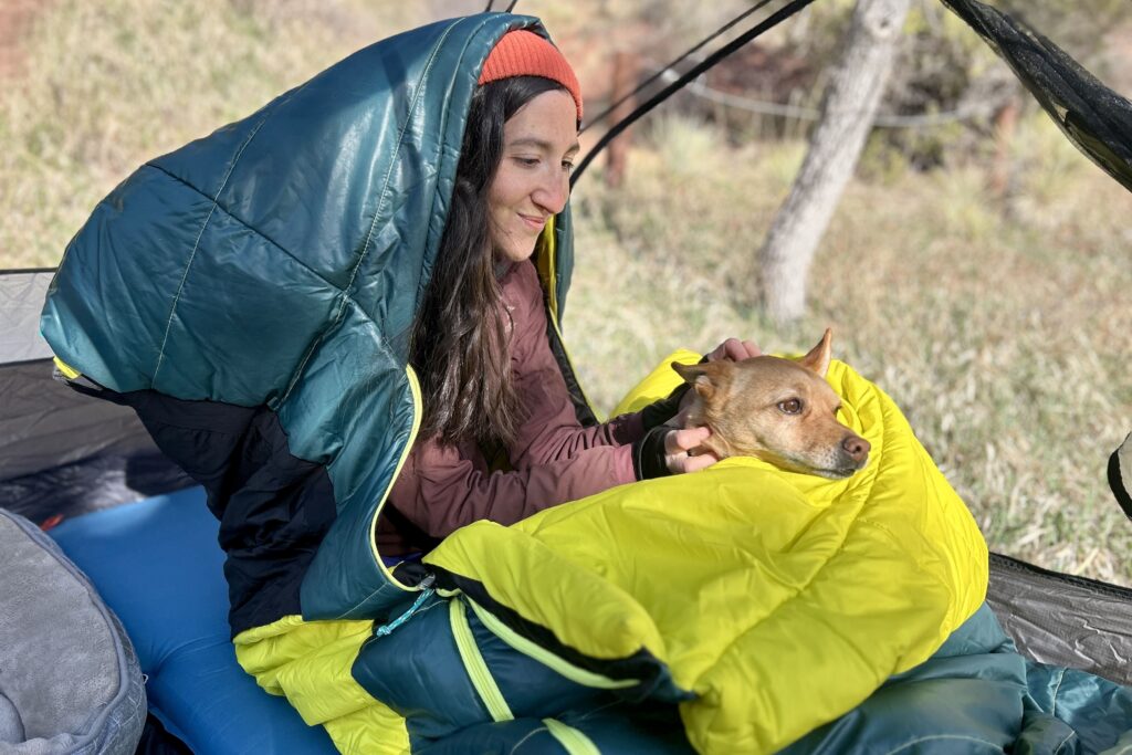 A woman sits up in a sleeping bag in a tent with her small dog in her lap.