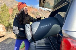 A woman puts a large, rolled-up camping mattress into the back of a pickup truck.