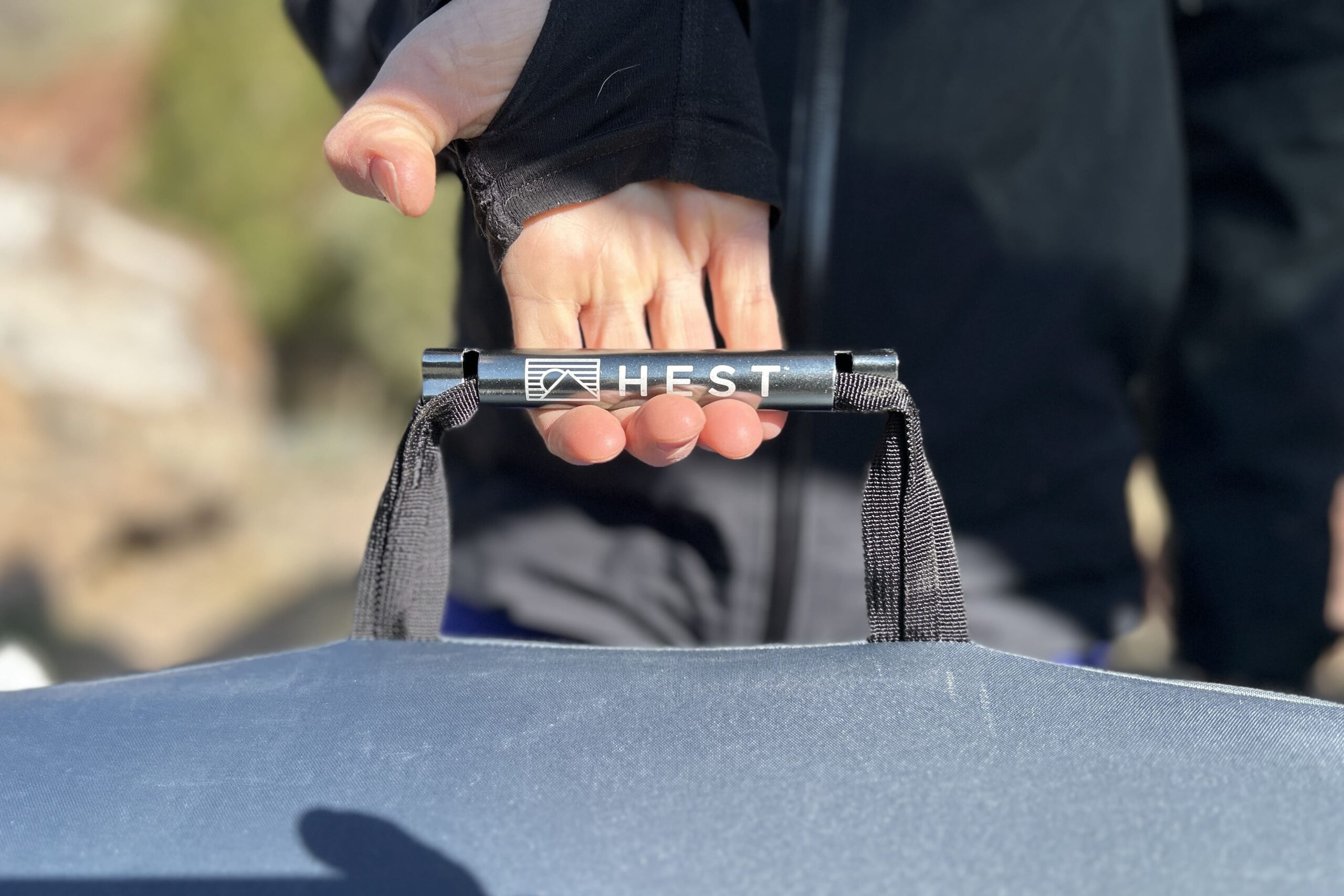 Close up image of a person's hand holding the thick nylon and metal handle of a camping mattress with the product label, HEST, prominently displayed.