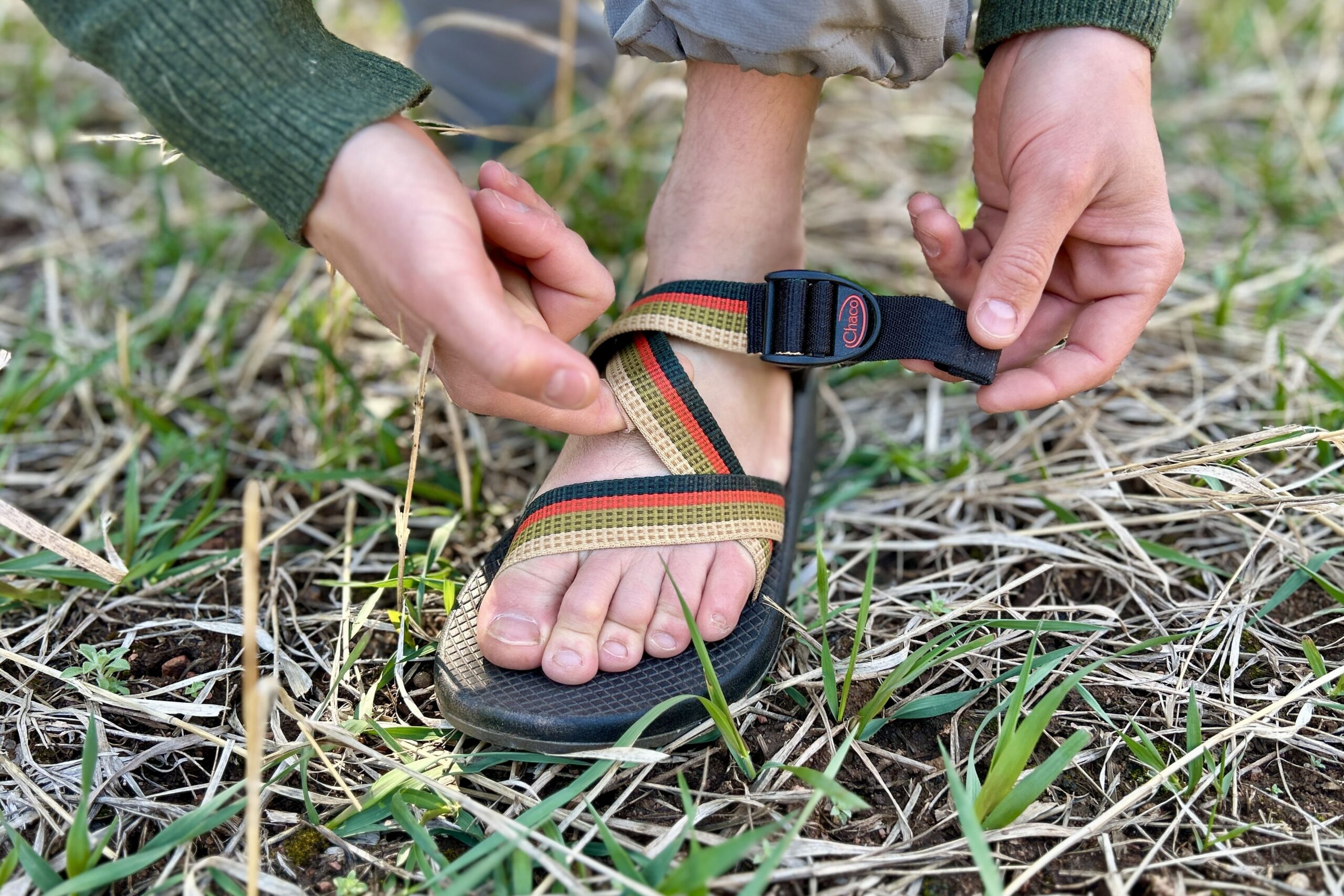 Close up image of a person adjusting a pair of hiking sandals in a grassy setting