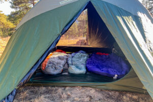 A tent full of camping gear is sitting in a filed. The doors of the tent are open on both sides so you can see straight through the tent.
