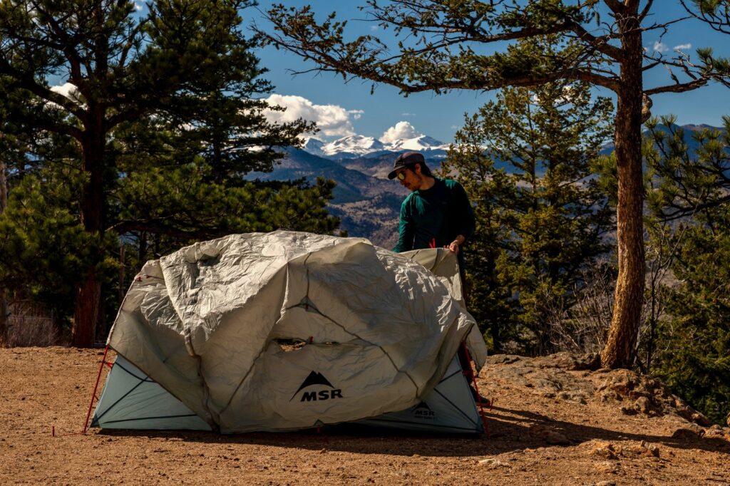 a hiker drapes the fly of a tent over the tent body with mountains and trees in the background.