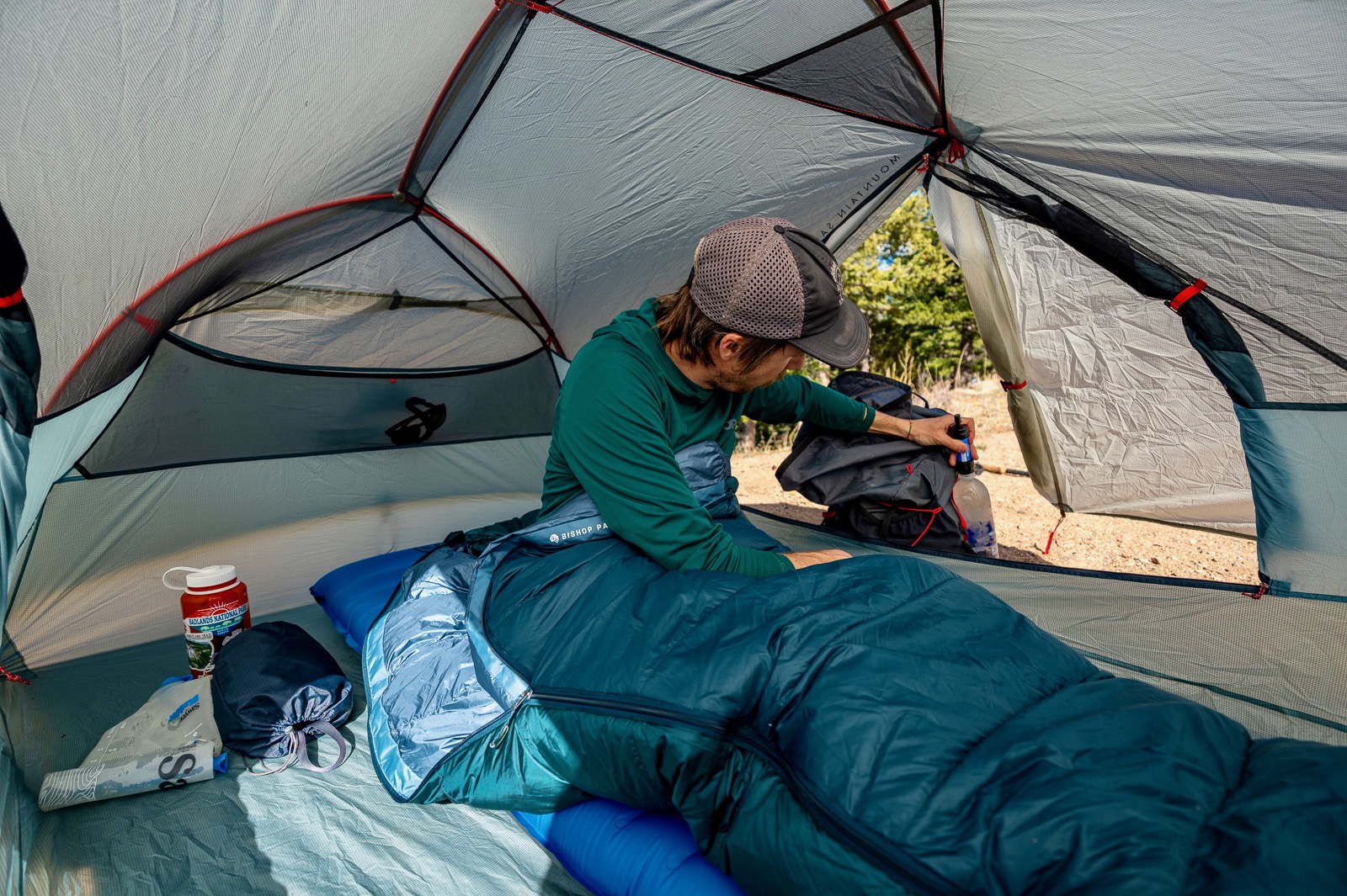 a hiker sits in a tent in a sleeping bag while he reaches for his water bottle.
