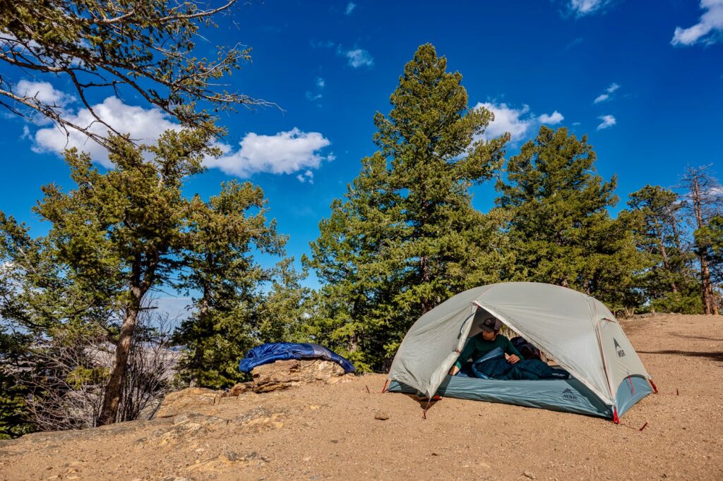An msr tent is pitched on dirt while a sleeping bag dries on a nearby rock. a hiker is in the tent and trees are in the background.
