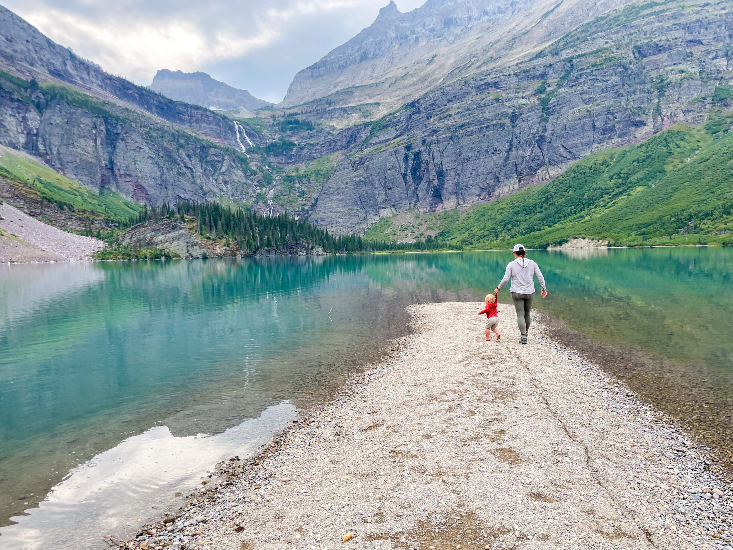 a mom and kid walk along a sandbar in a lake