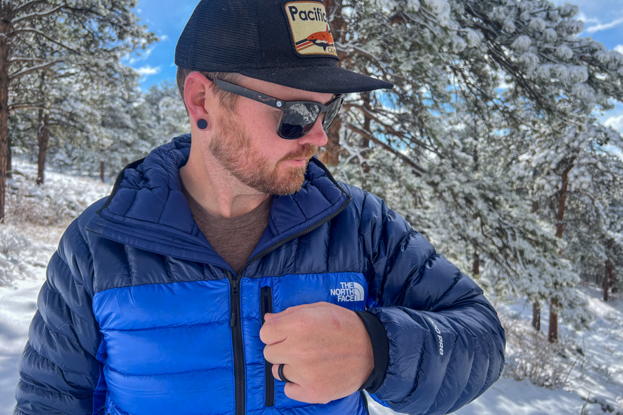 a man standing in a snowy forest. He's wearing a blue jacket and adjusting the zipper of a jacket pocket.