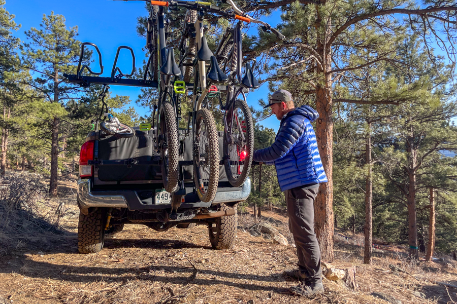 A man standing near a bike rack attached to a pickup truck. The man is in a forest surrounded by pinetrees.