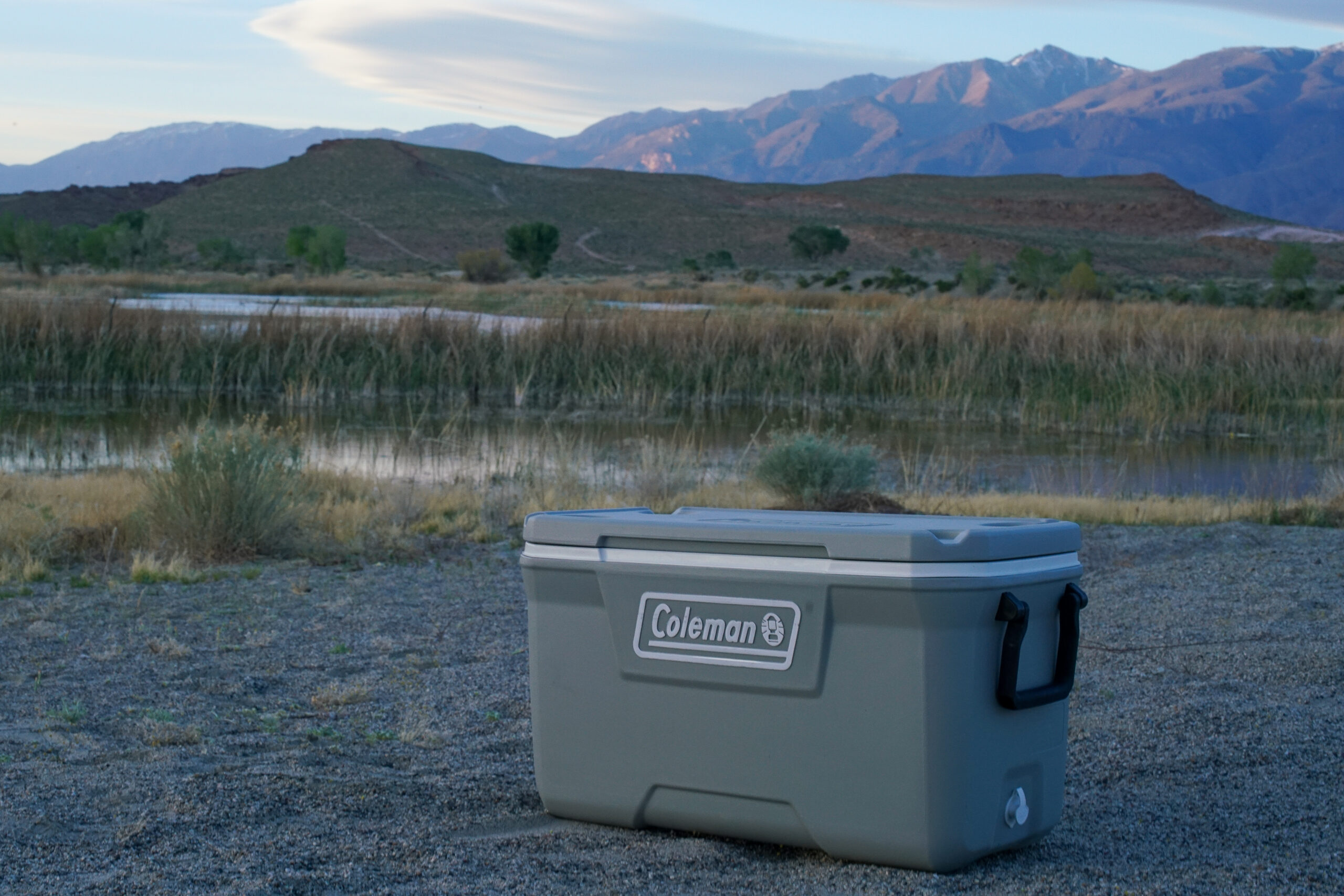 The Coleman 316 Series 70QT cooler sits on sand beside a body of water with mountains in the background.