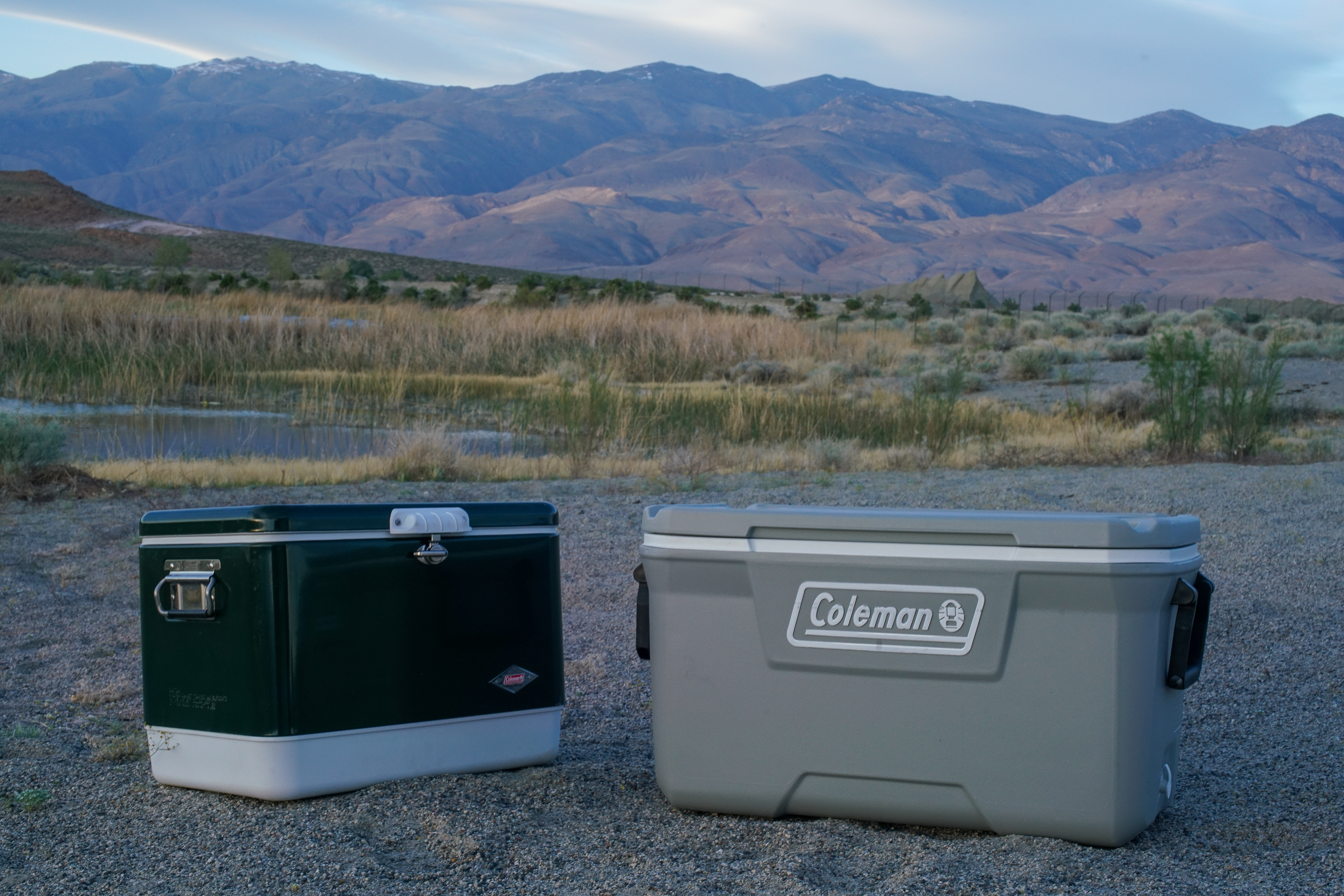 The Coleman Steel Belted cooler Coleman 316 Series 70QT cooler sit beside each other on sandy ground with mountains in the background.