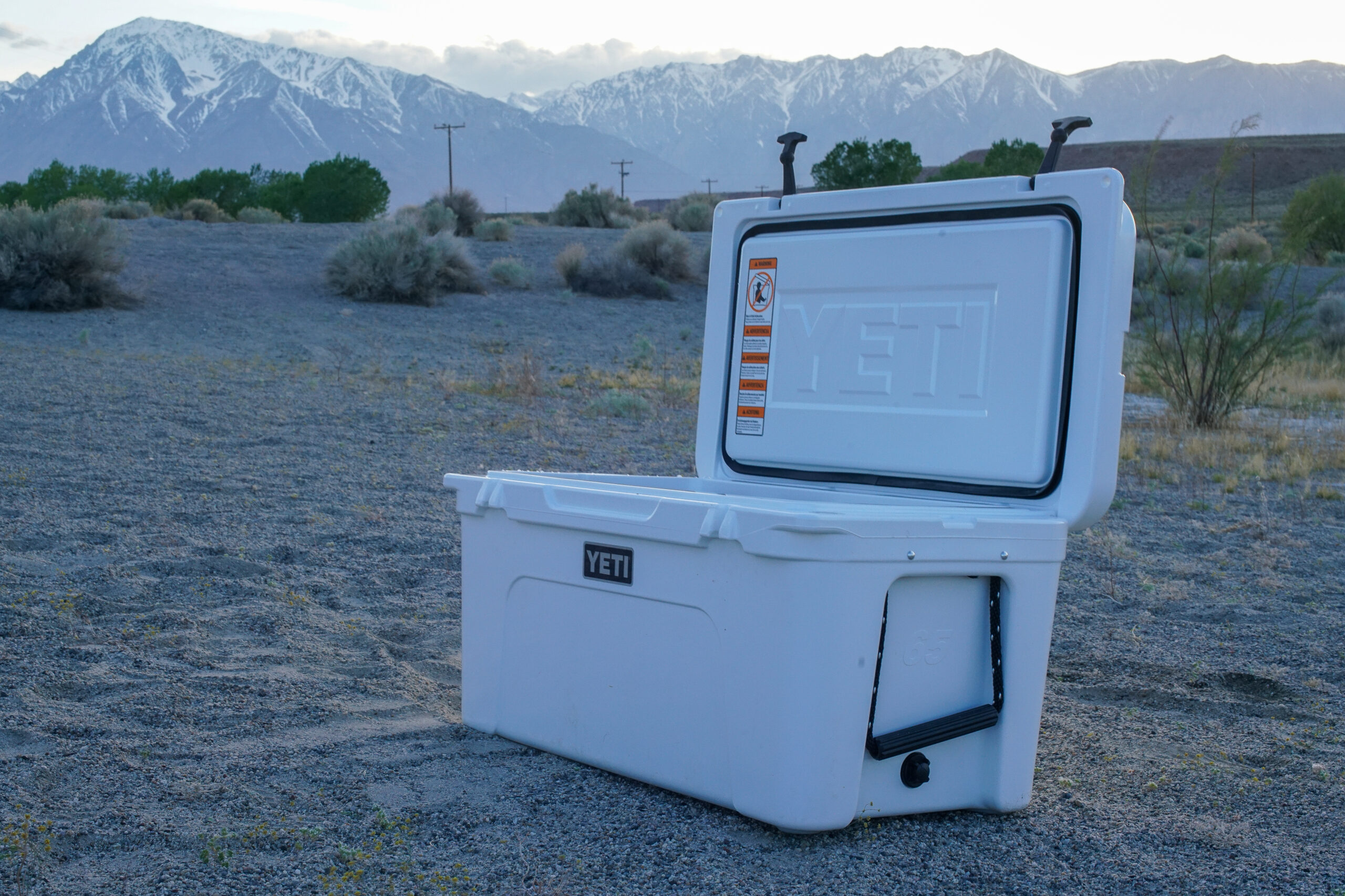 The YETI Tundra 65 cooler sits on sandy ground with its lid open and mountains in the background.