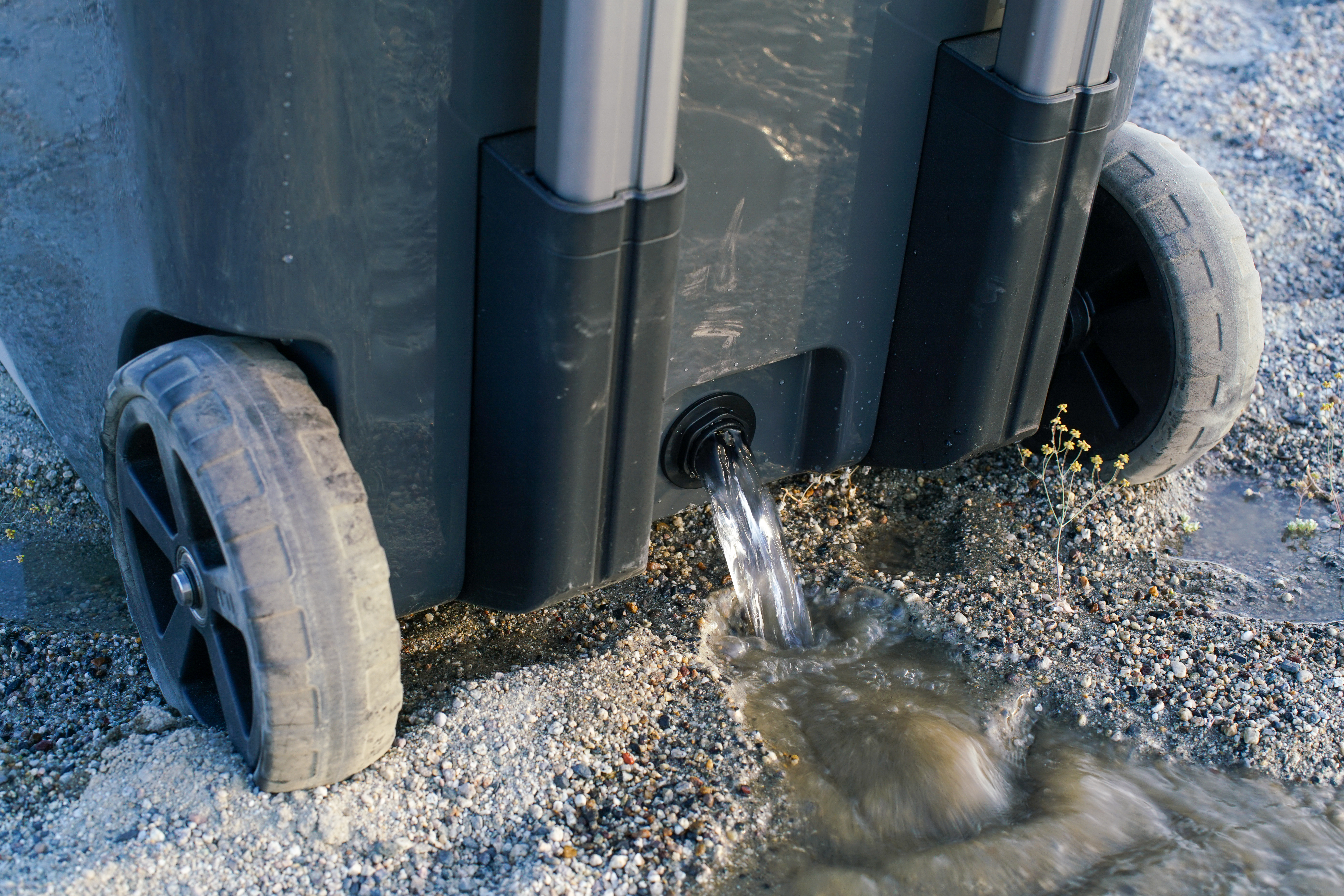 A close up of water pouring out of the YETI Roadie 48 cooler's drainage hole onto sandy ground.