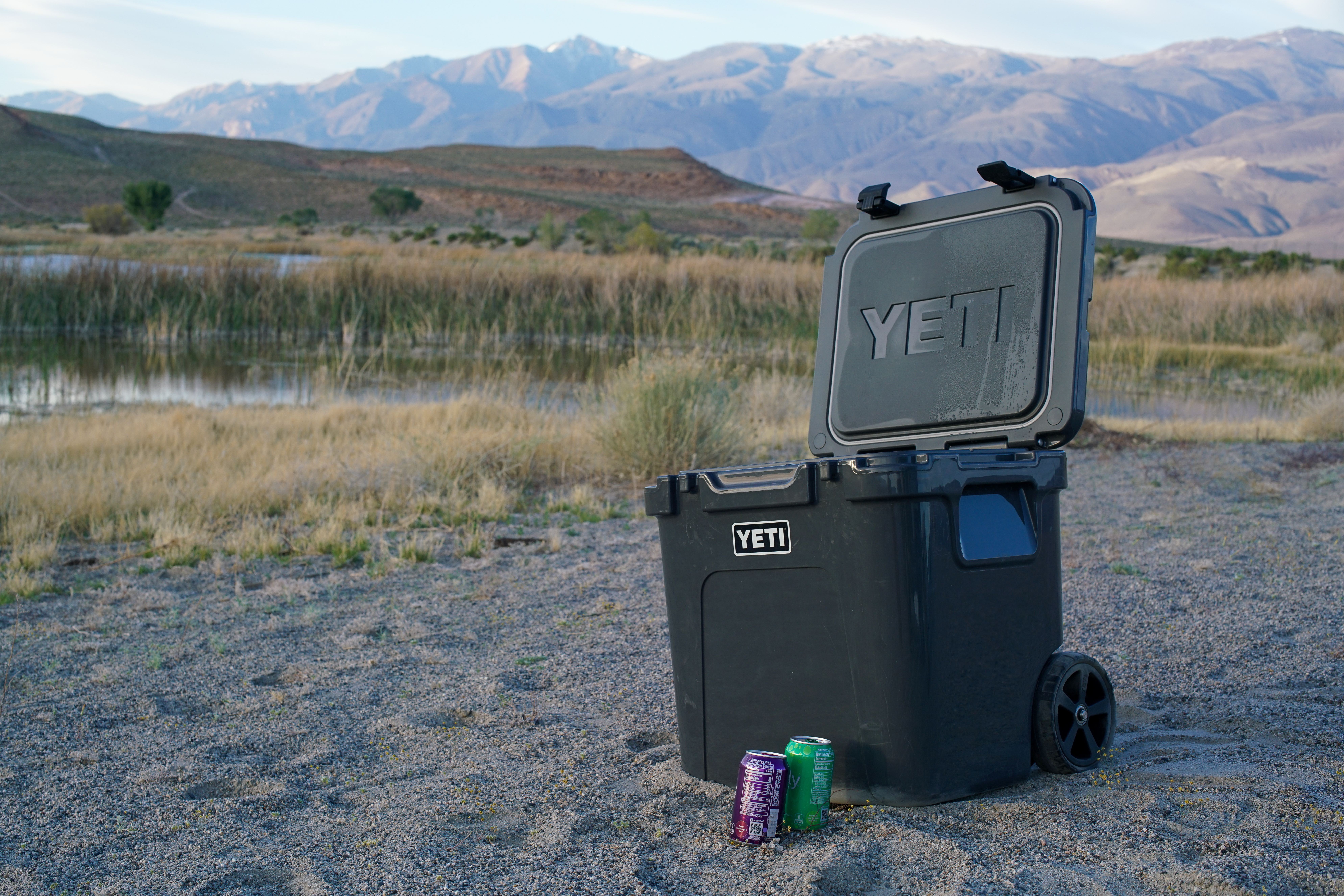 The YETI Roadie 48 cooler sits on sandy ground with its lid open. Two canned drinks sit beside it.