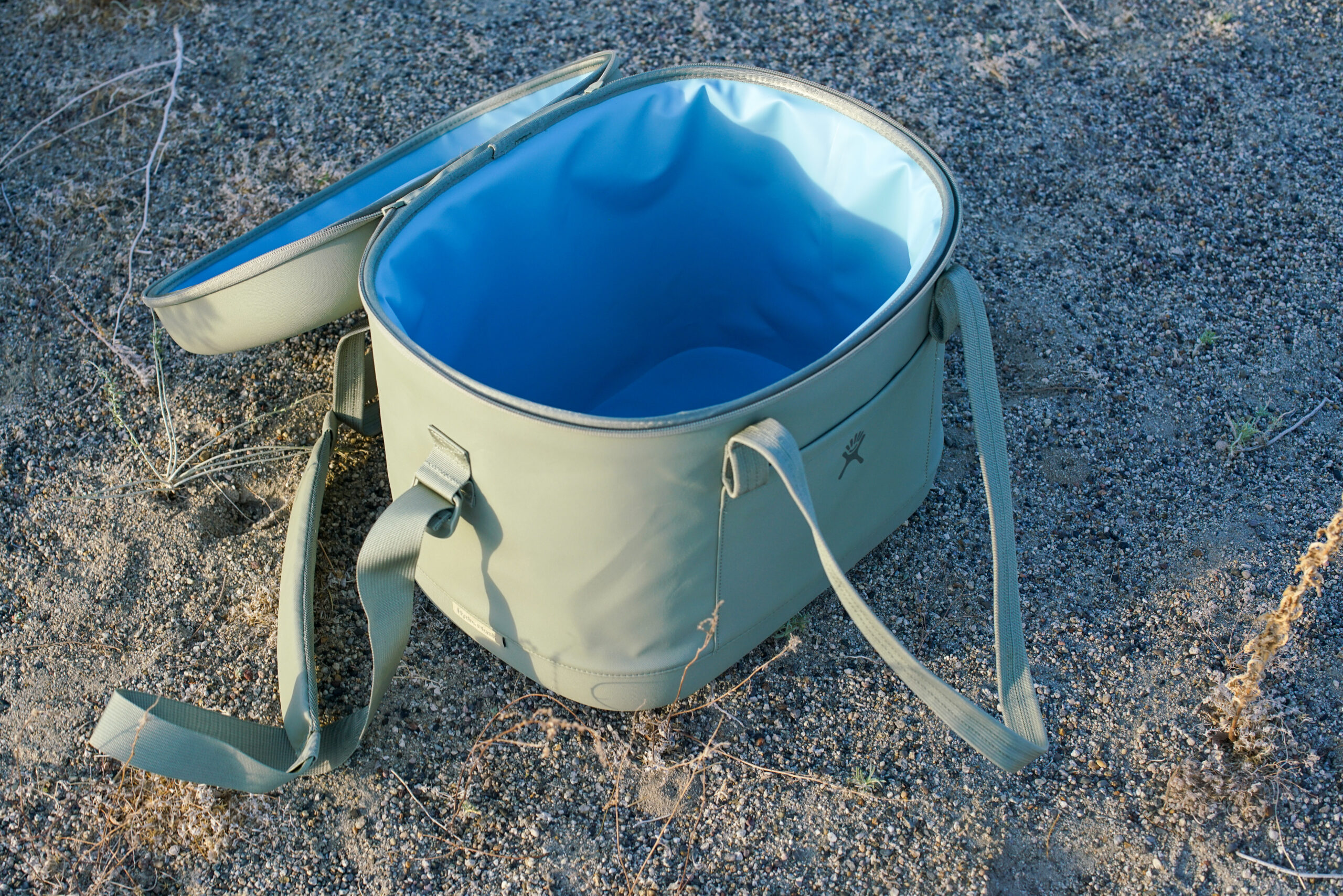 An open Hydro Flask Carry Out 20L cooler sits on sandy ground. The cooler is empty.