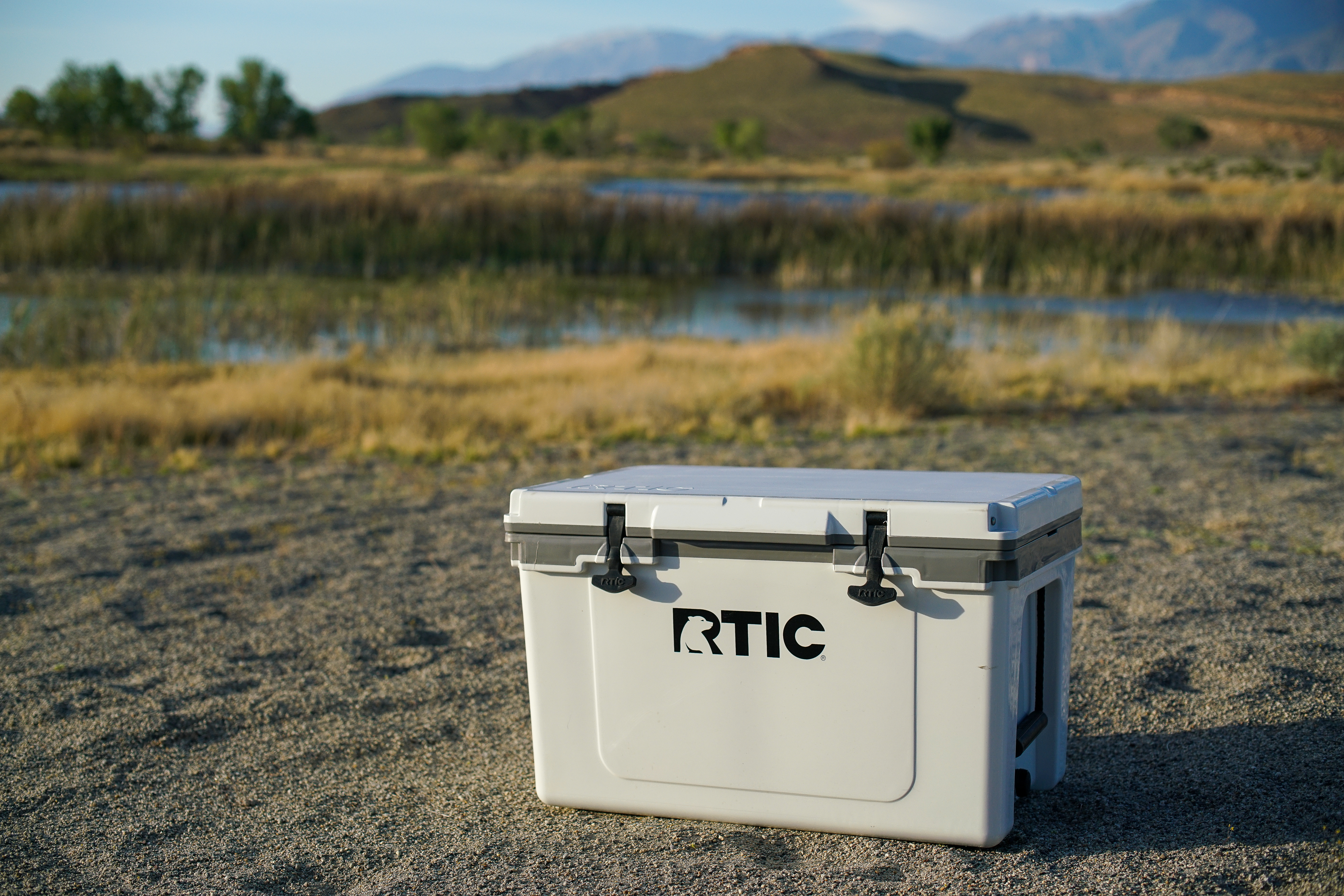 The RTIC Ultralight 52 cooler sits on sandy ground with a body of water and mountains in the background.
