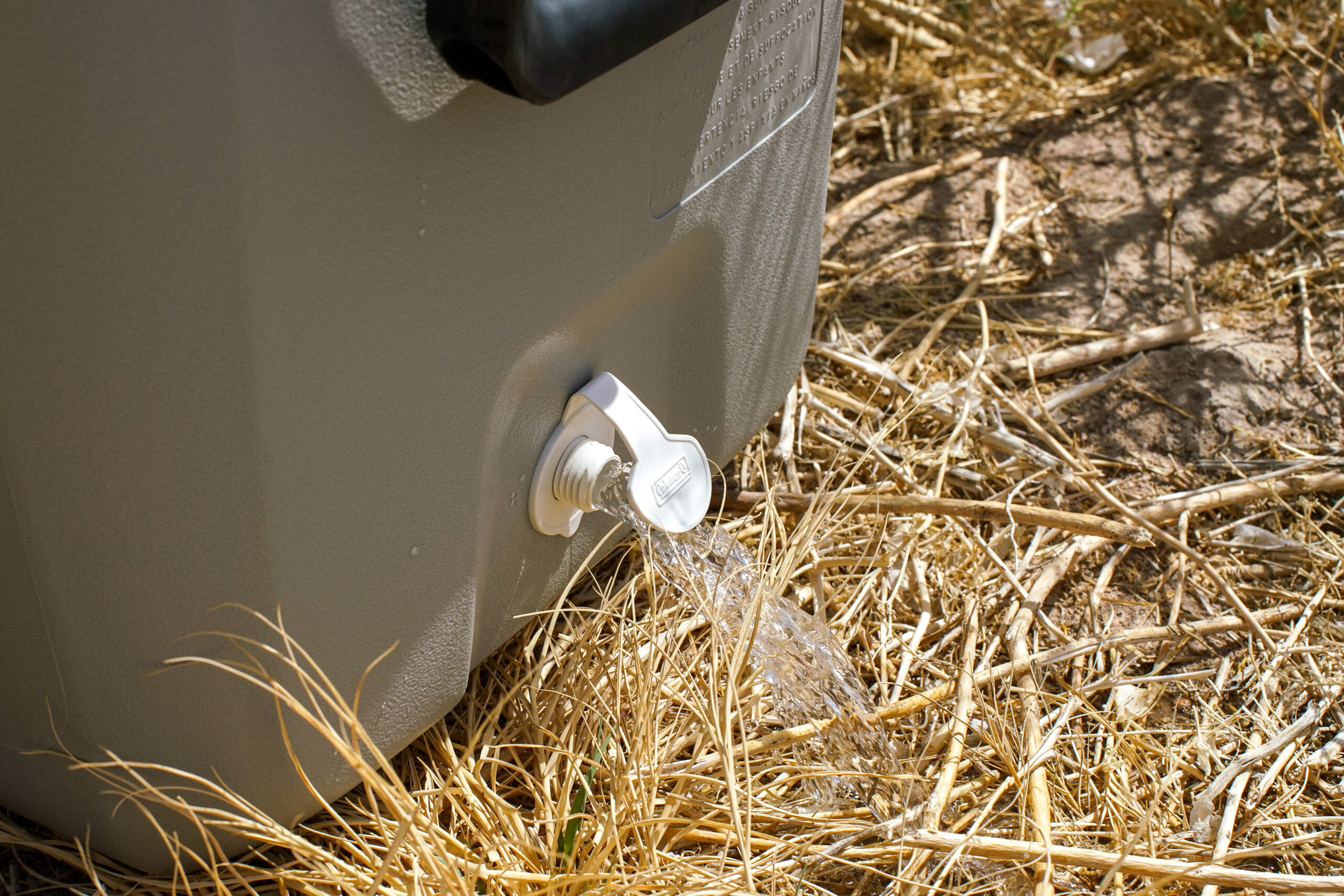 A close up of the drainage hole on the Coleman 316 Series 70QT cooler with water spilling onto straw on the ground.
