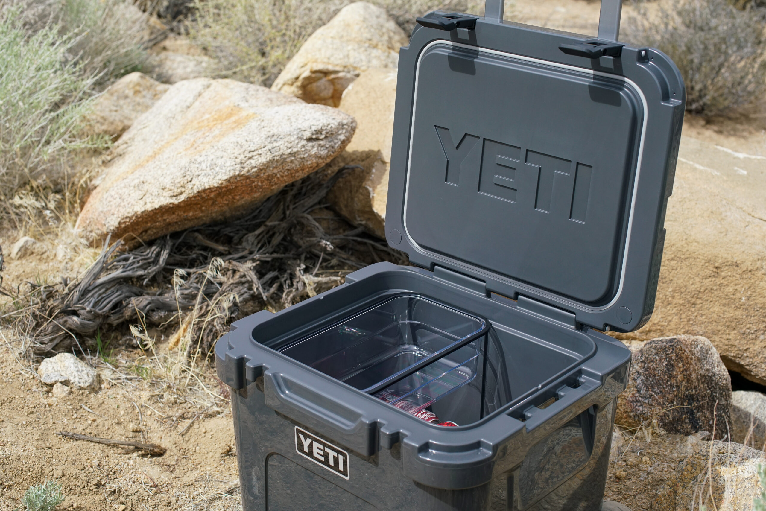 The YETI Roadie 48 sits in a rocky environment. The cooler is open and a clear plastic basket sitting in the cooler is visible.