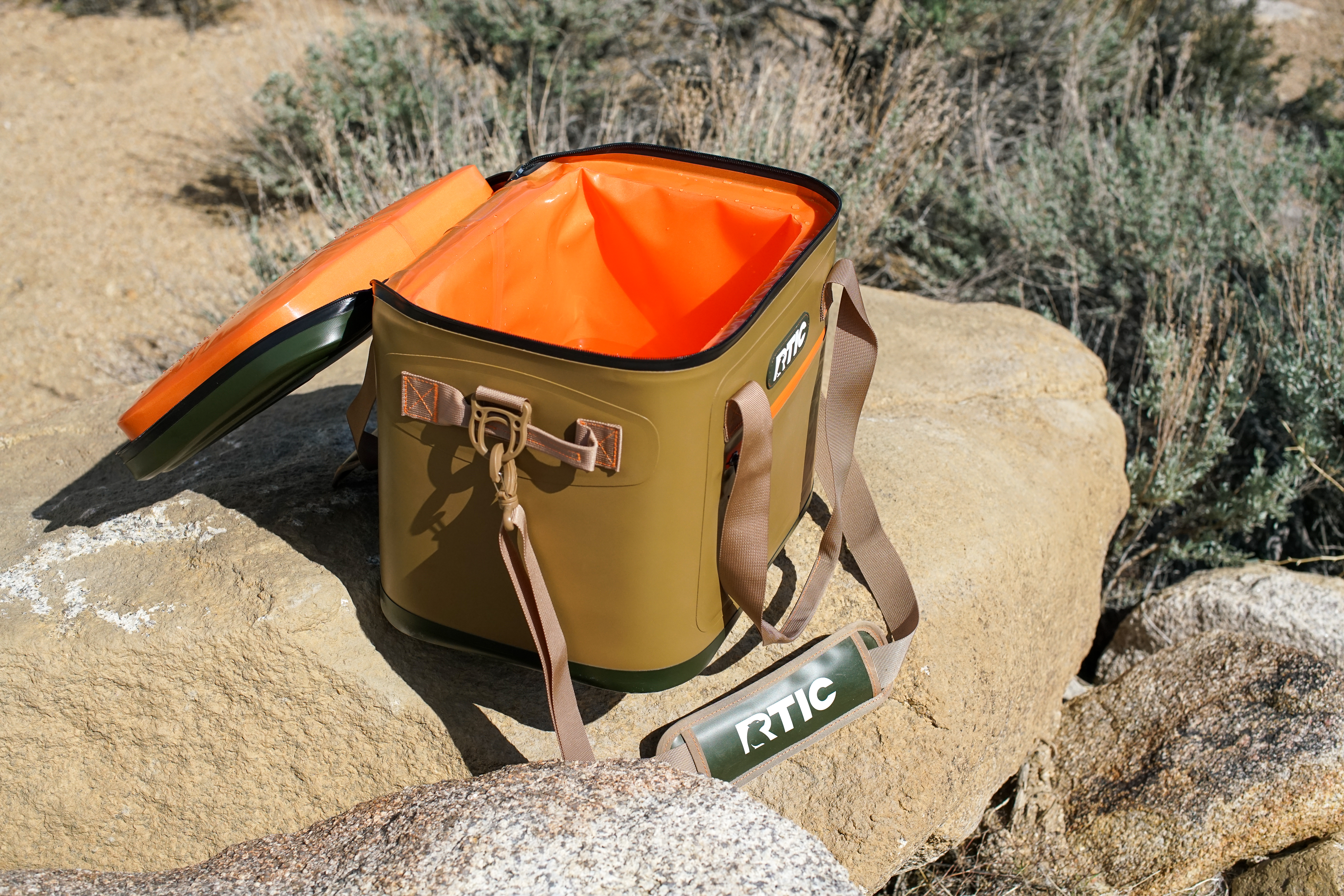 The RTIC Soft Pack cooler sits on a rock. The lid of the cooler is open, revealing an empty interior.