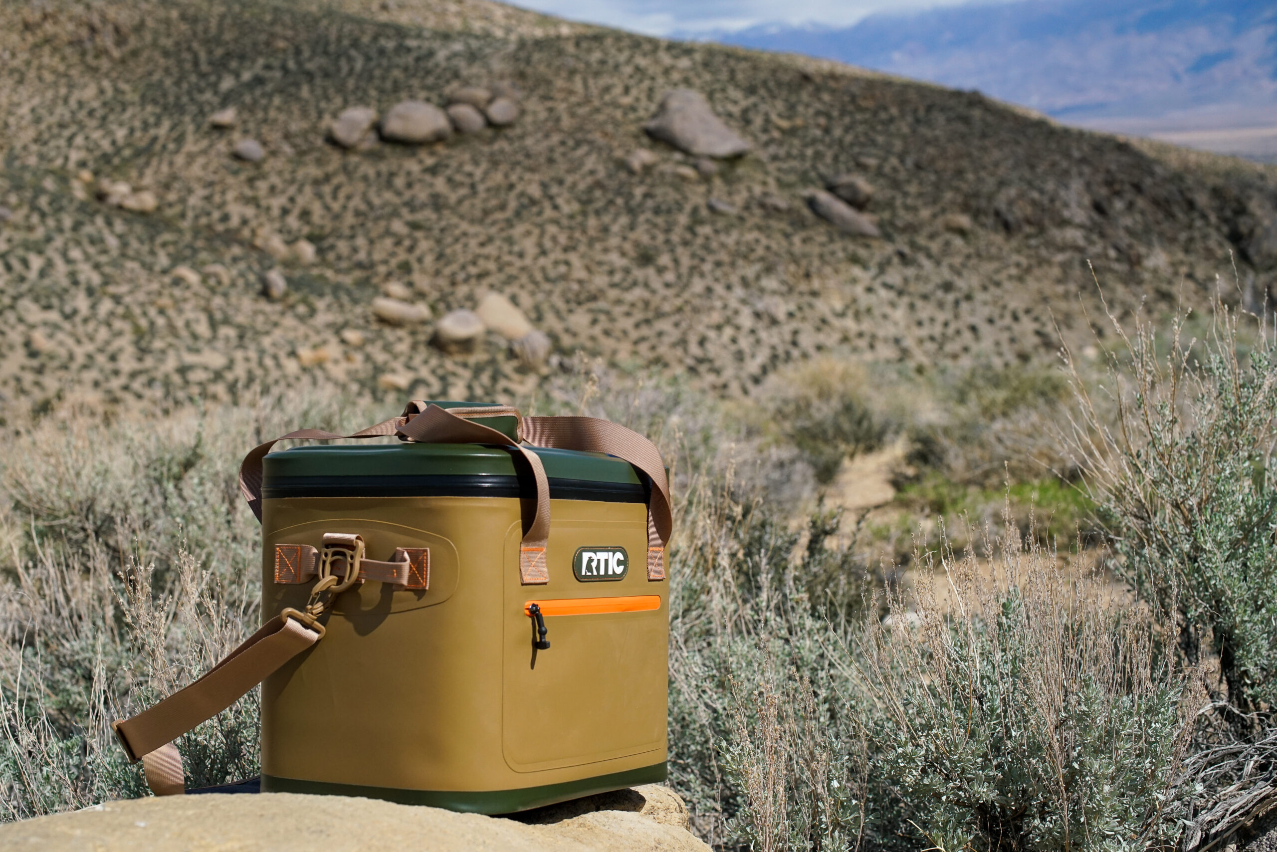 The RTIC Soft Pack cooler sits on a rock with sage brush and boulders in the background.