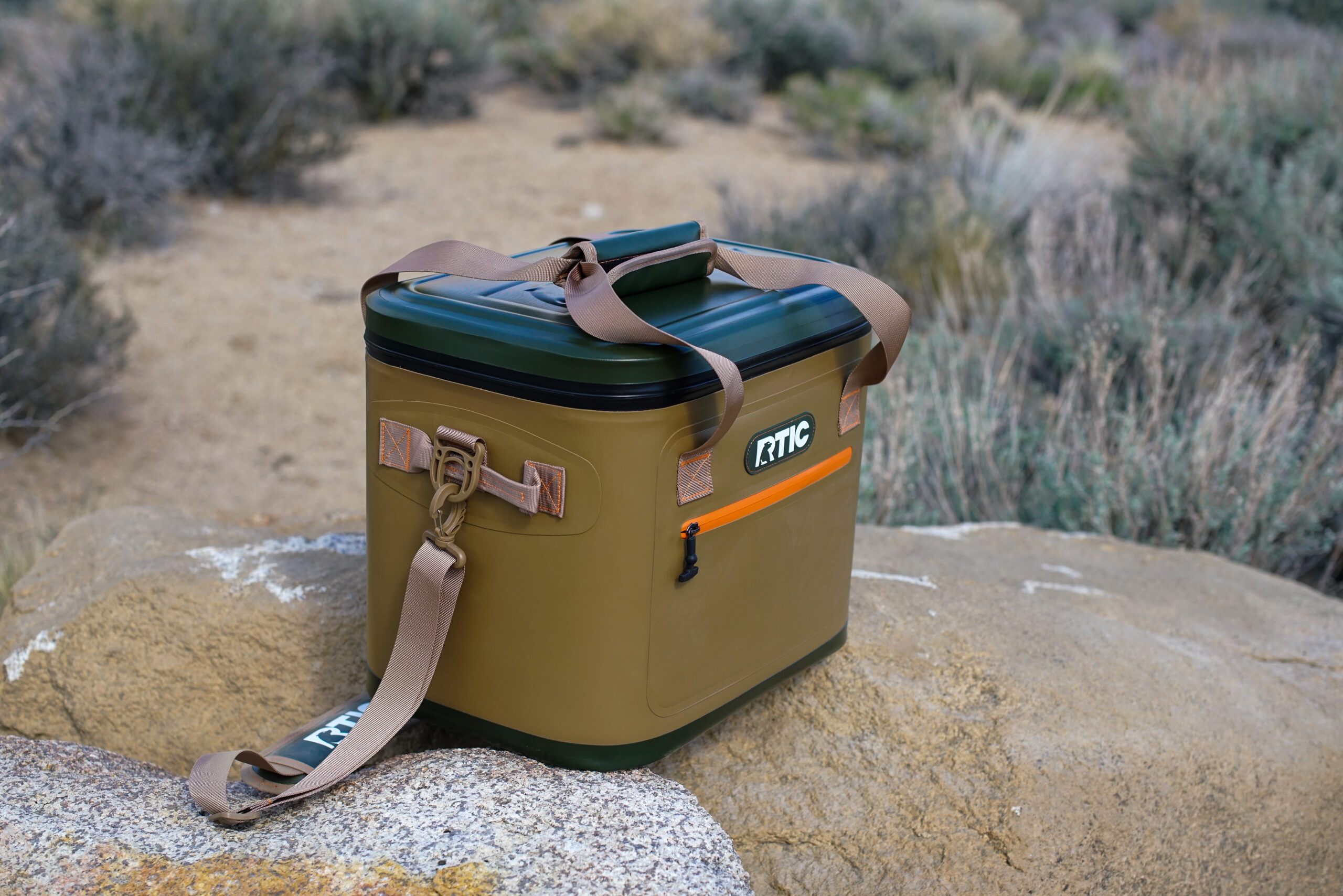 The RTIC Soft Pack cooler sits on a rock with sage brush in the background.