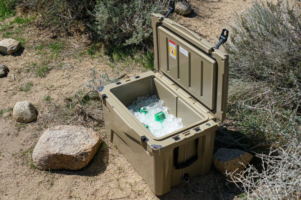 The XSpec 60QT cooler sits on the ground with its lid open, revealing a layer of ice cubes along with several canned beverages.
