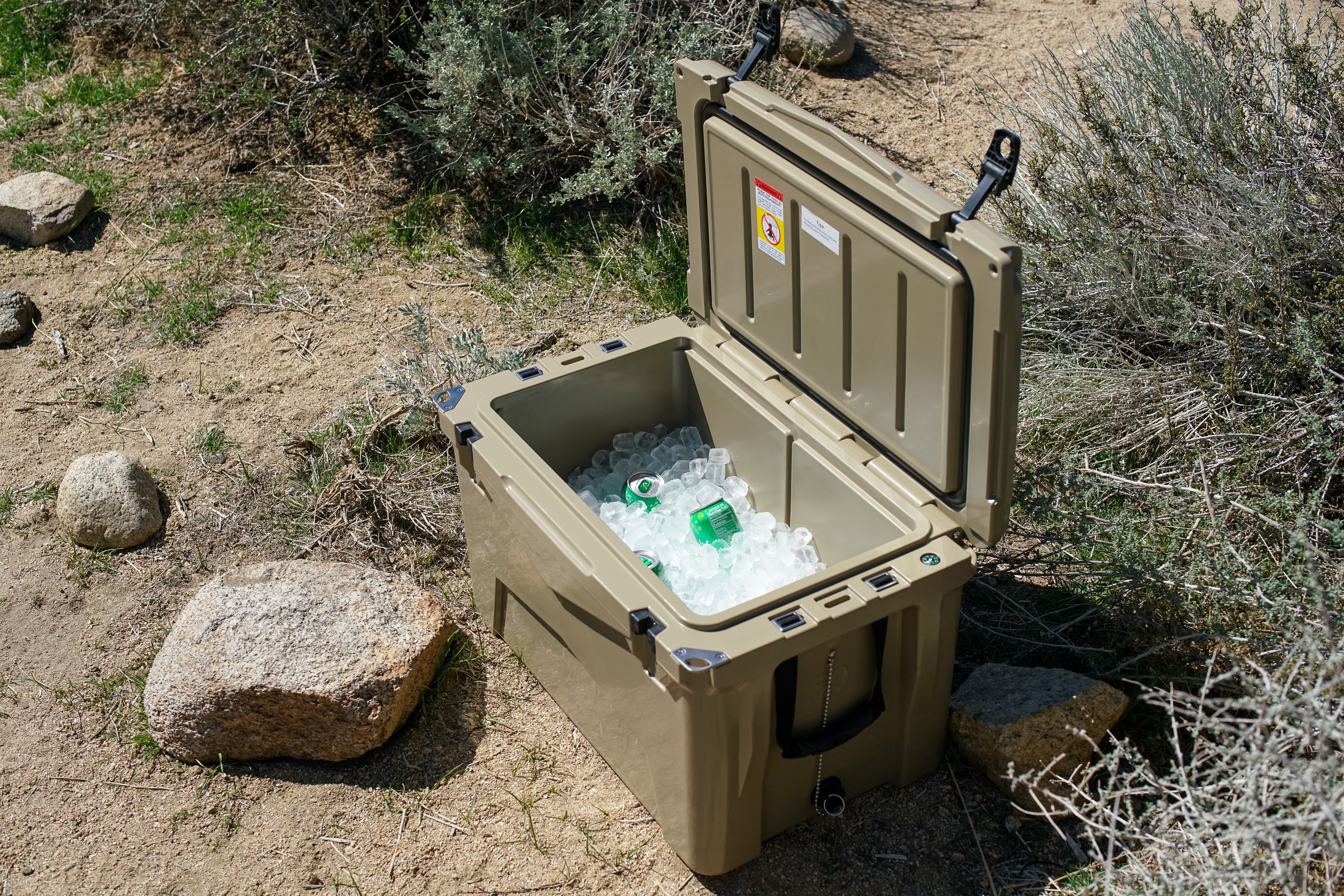 The XSpec 60QT cooler sits on the ground with its lid open, revealing a layer of ice cubes along with several canned beverages.