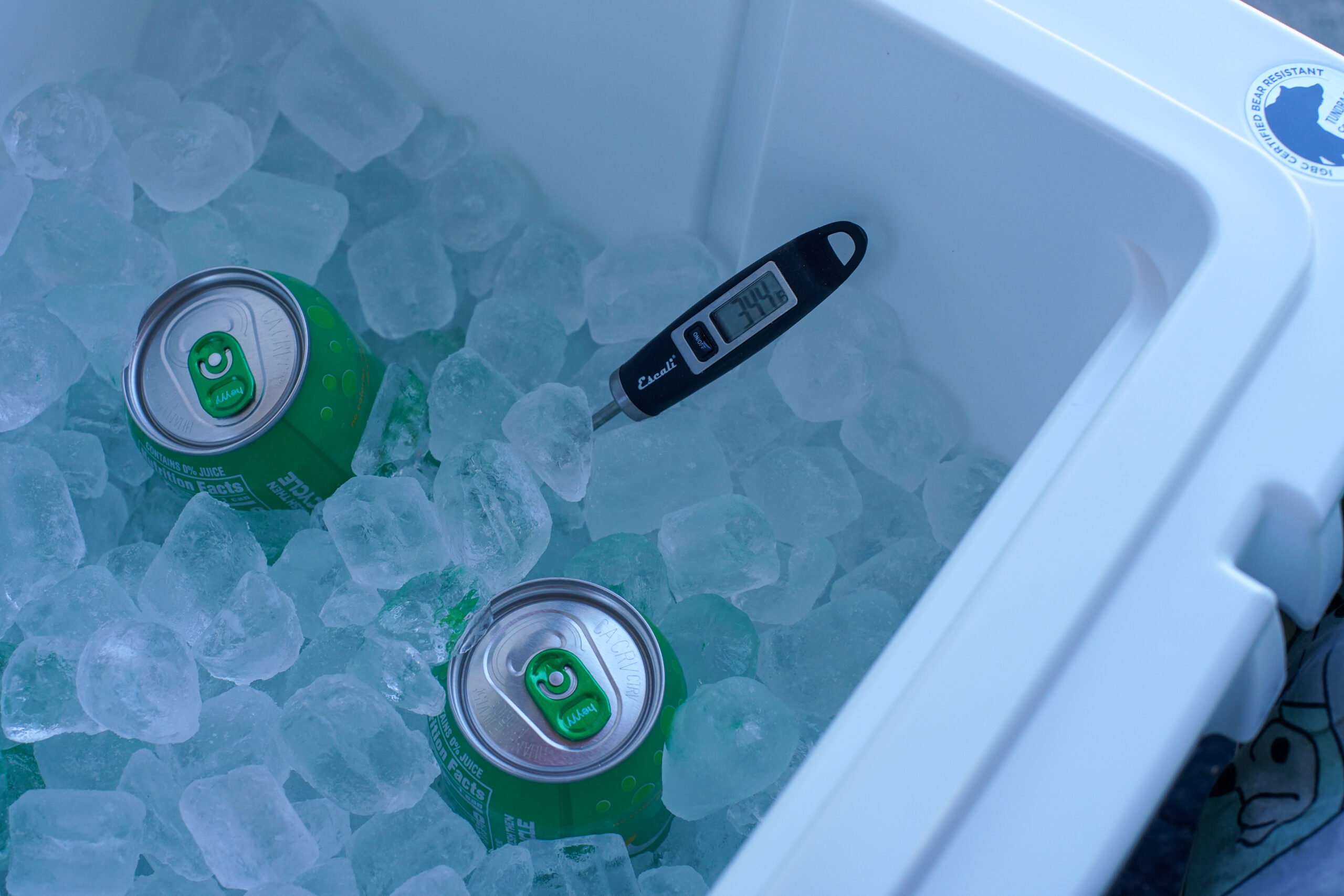 A thermometer reading 34.4 degrees fahrenheit sticks out of a bed of ice in the corner of the YETI Tundra 65 cooler. Two canned drinks poke out of the ice.