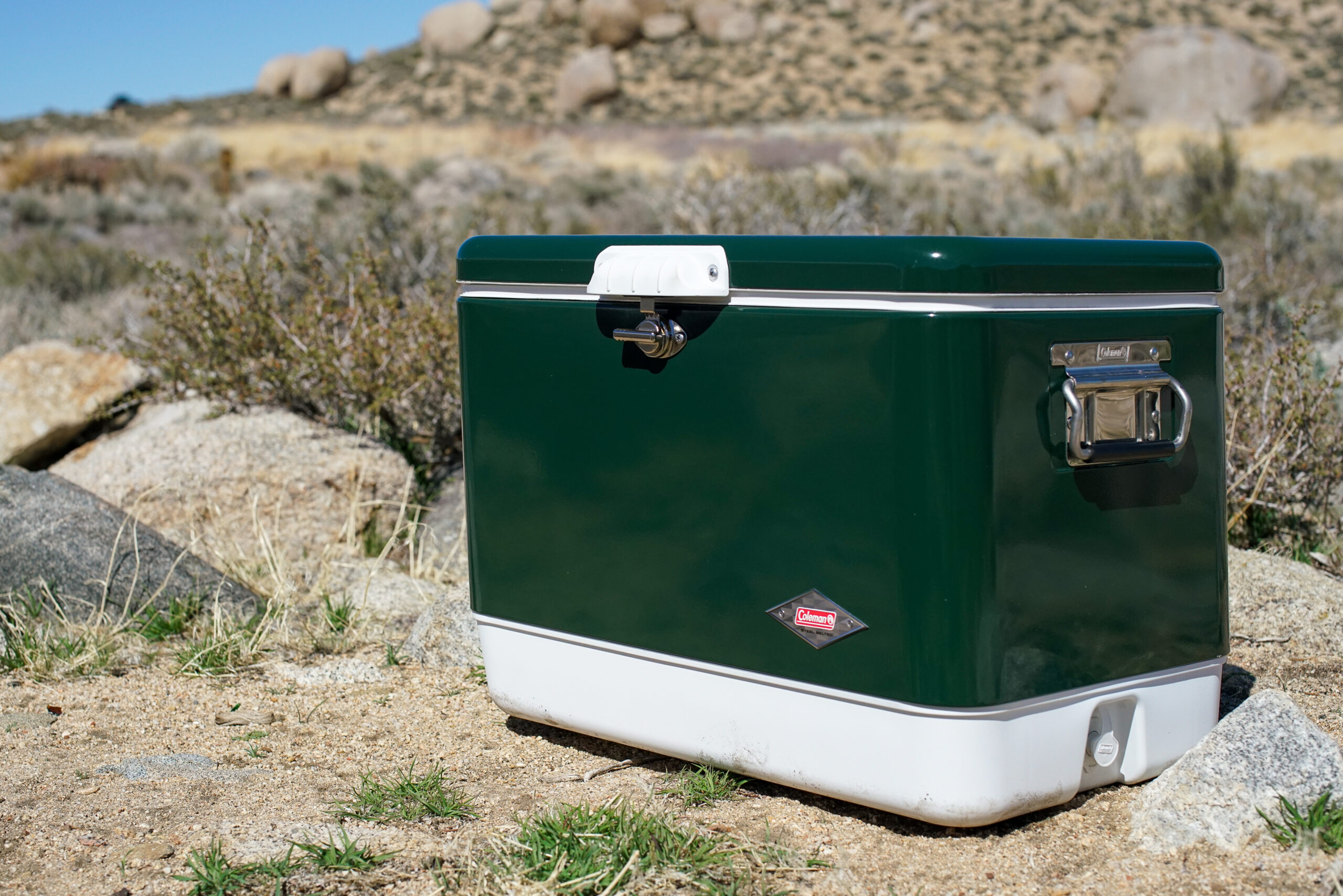 The Coleman Steel Belted cooler sits on the ground with boulders in the background.