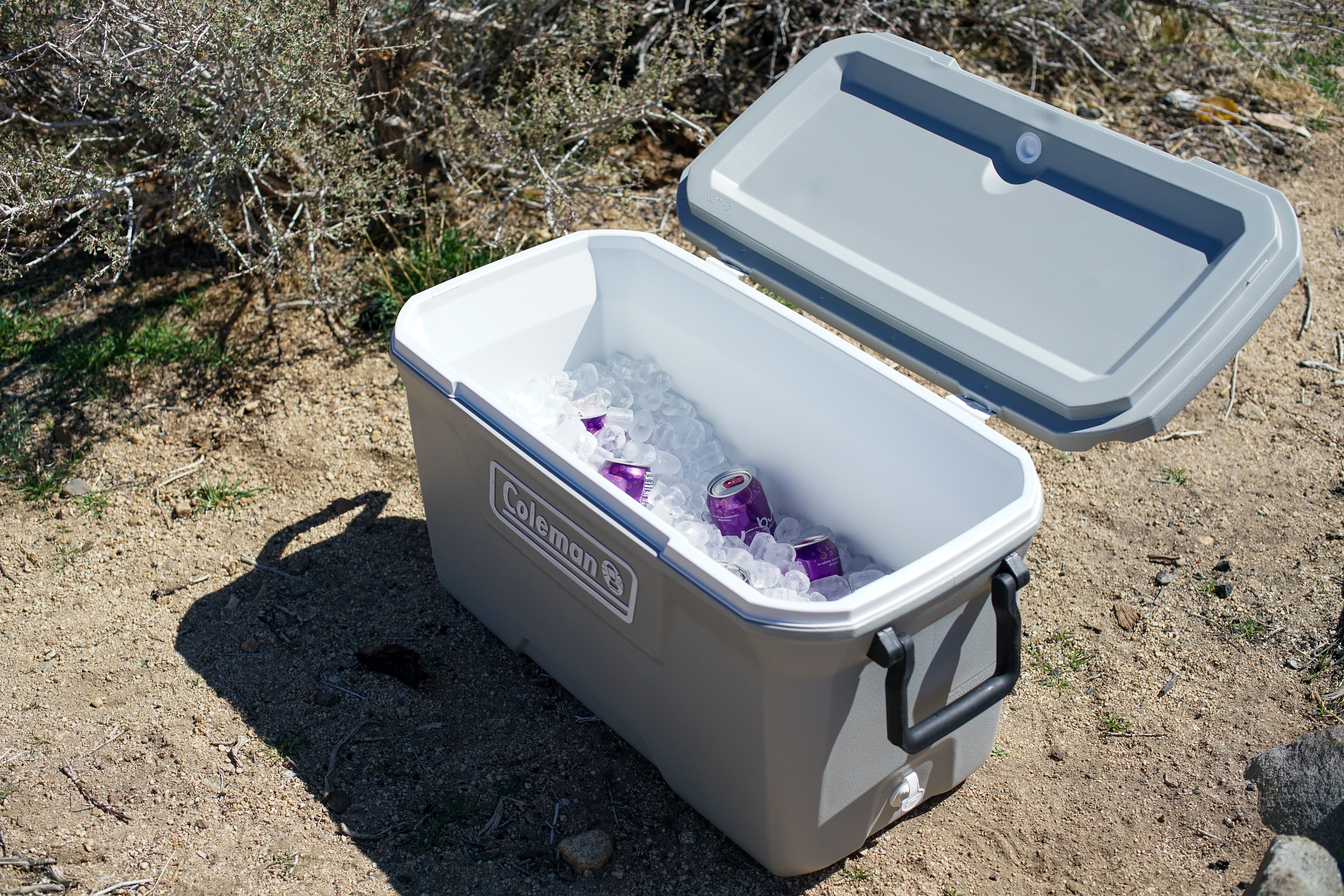 The Coleman 316 Series 70QT cooler sits on the ground with its lid open. The cooler is full of ice and some canned drinks are visible.