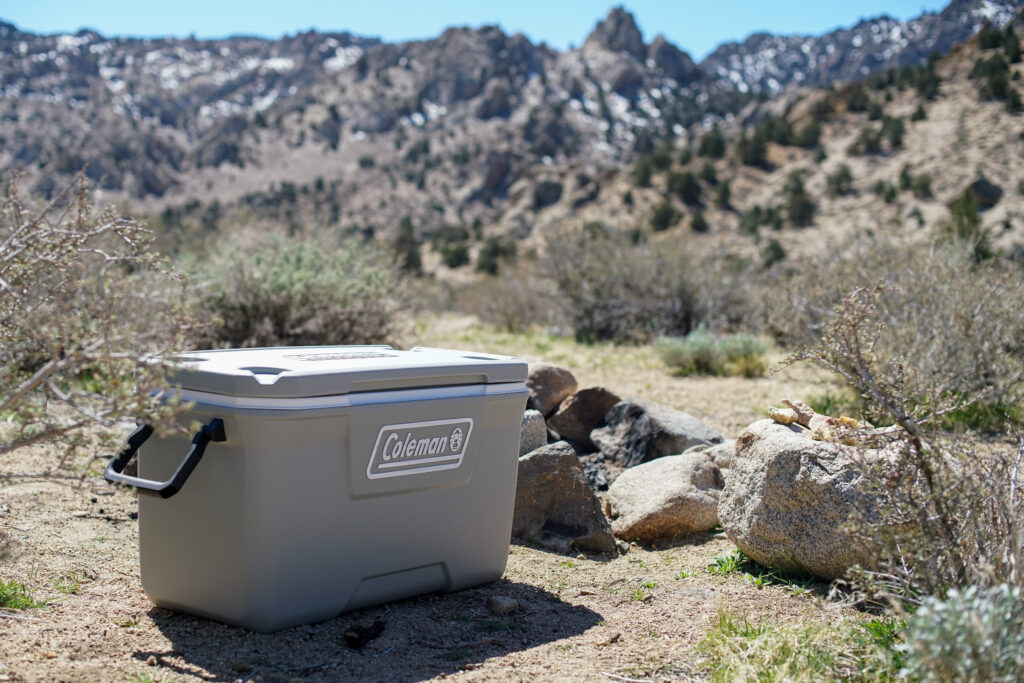 The Coleman 316 Series 70QT cooler sits on rocky terrain with mountains in the background.