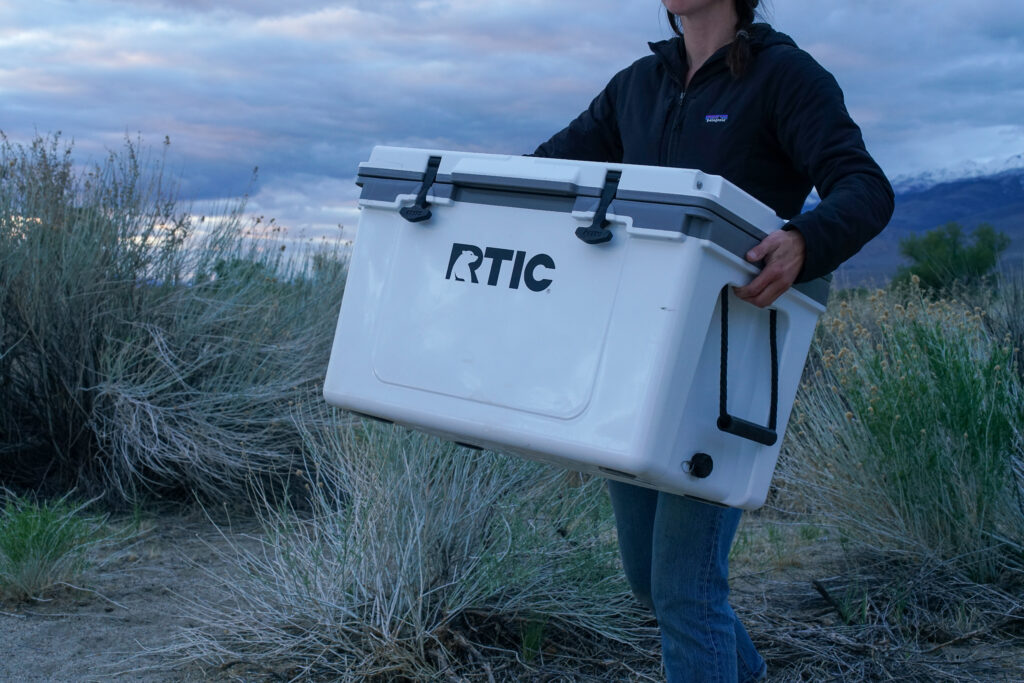 A close up of a person carrying the RTIC Ultralight 52 cooler by its built-in molded side handles.