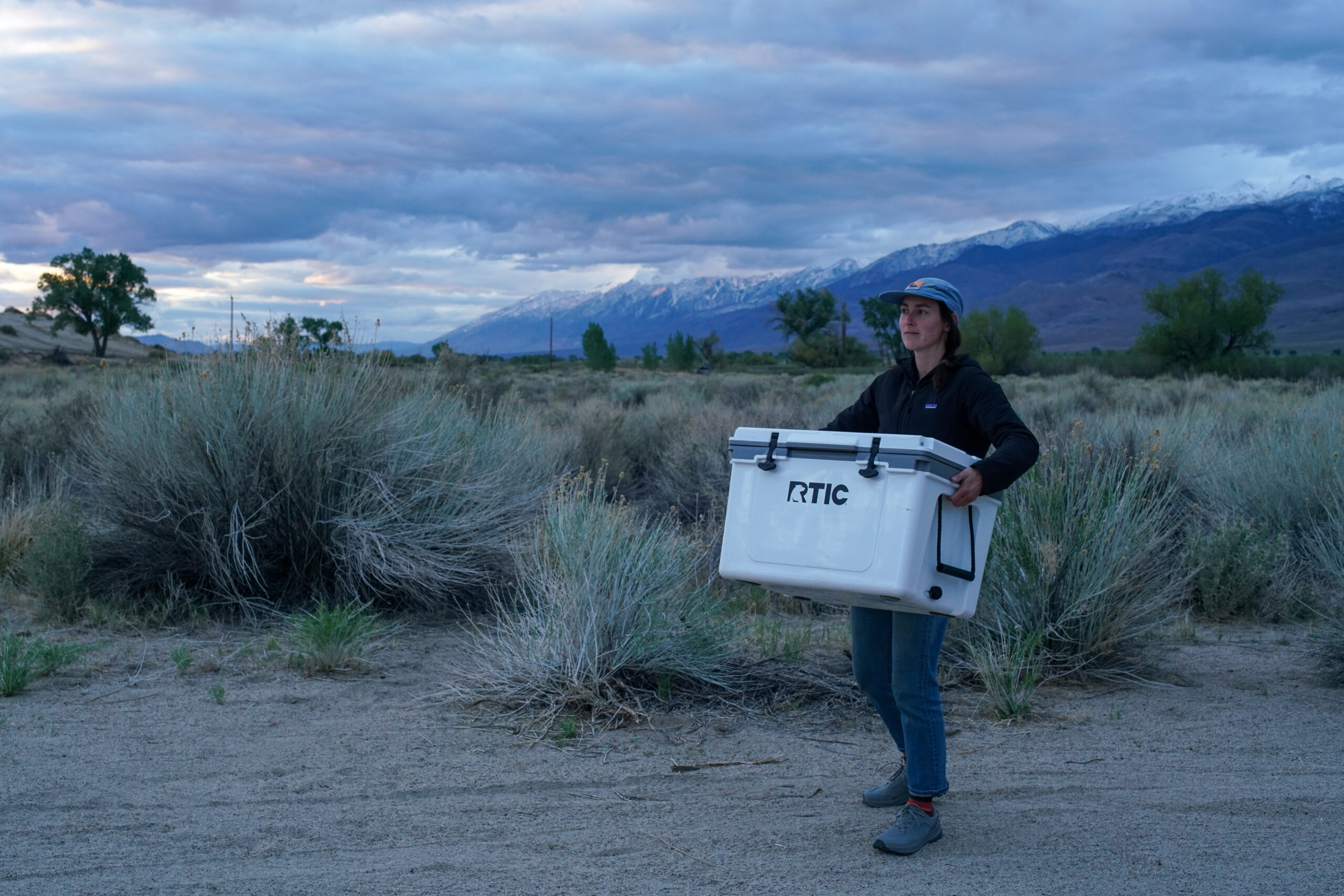A person carries the RTIC Ultralight 52 cooler by the molded side handles with mountains and sage brush in the background.