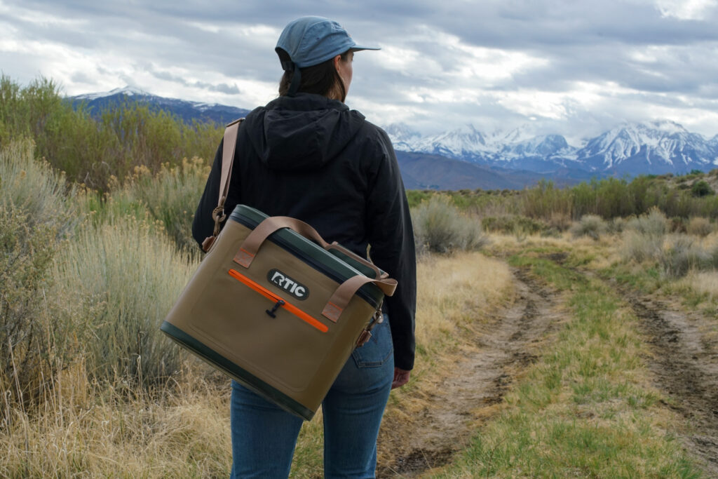 A person carries the RTIC Soft Pack cooler over their shoulder. Their back is turned towards the camera as they walk down a path with mountains in the distance.