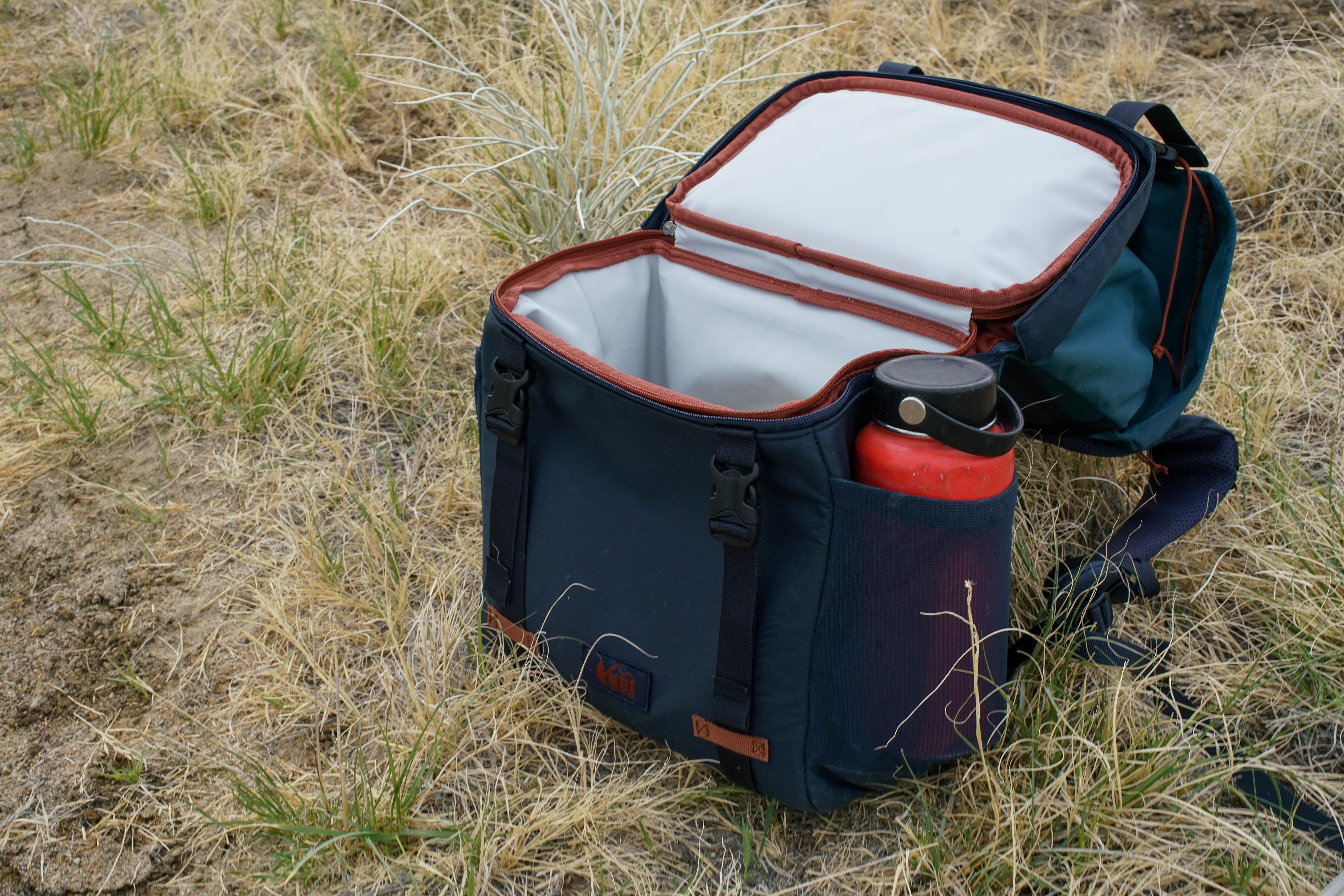 The REI Cool Trail Split Pack sits on grassy ground, unzipped at its middle to reveal the cooler built into the bottom half of the pack.