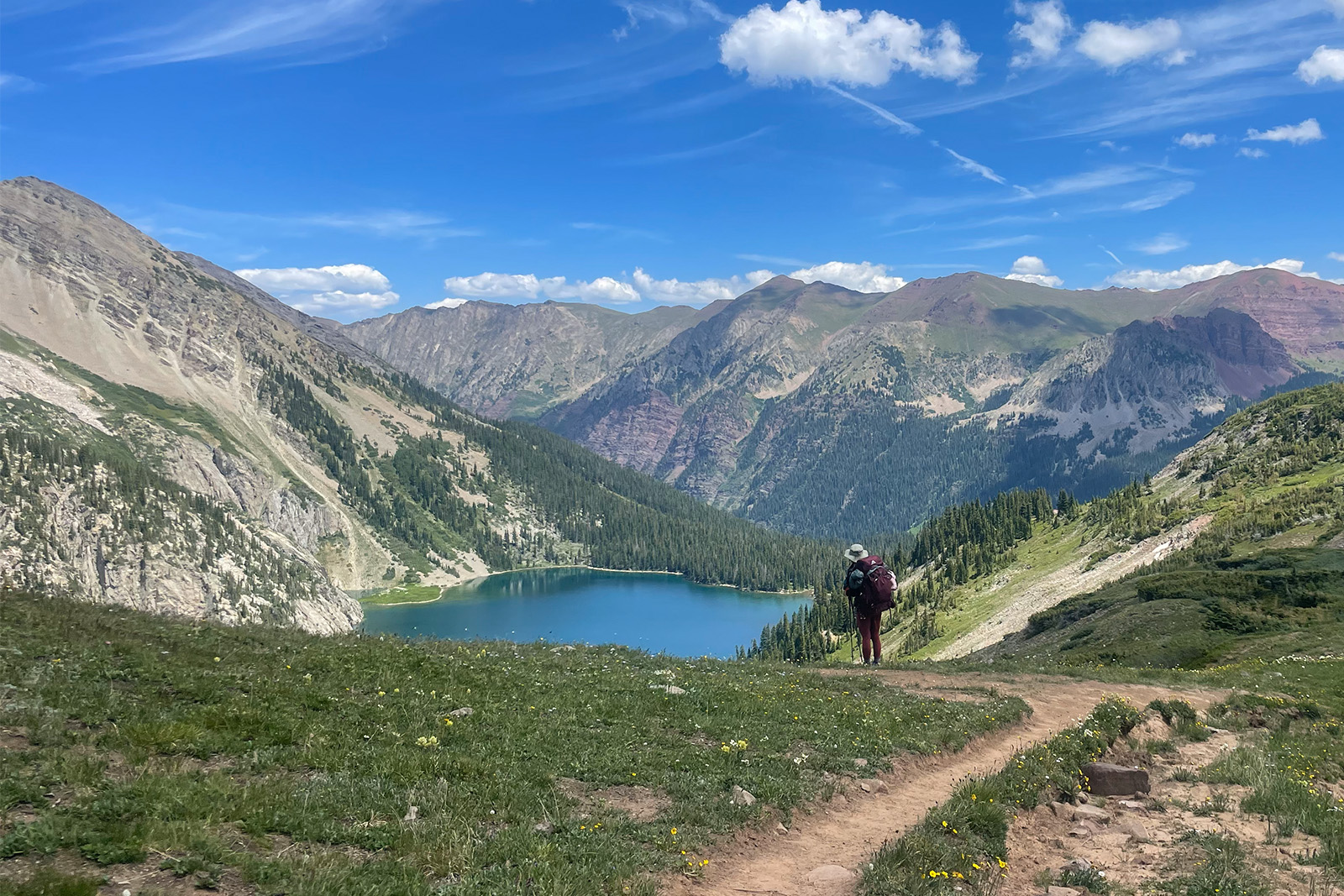 A person wearing a backpack is standing on a trail admiring a view of a lake. The lake is surrounded by mountains.