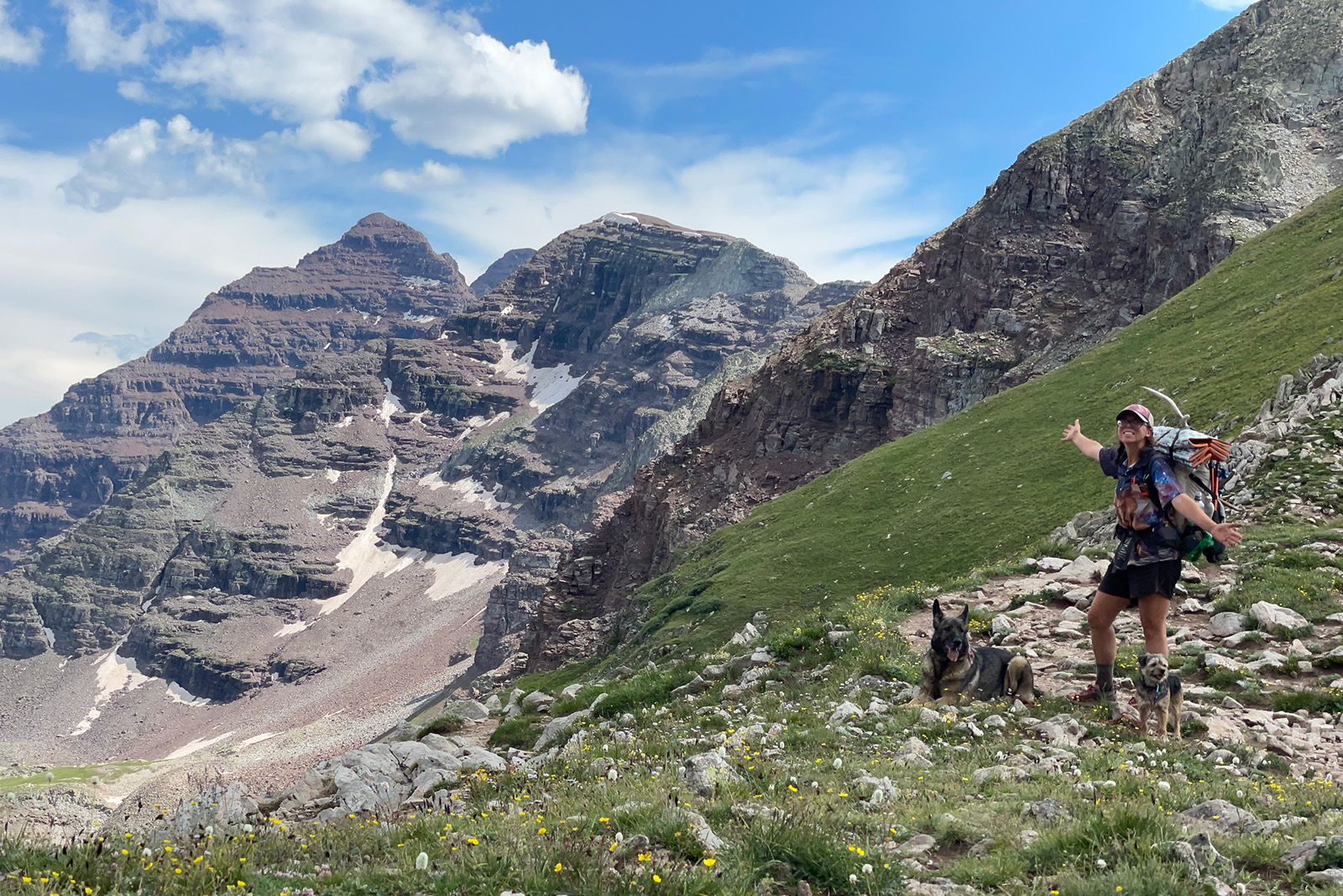A person wearing a backpack is standing next to two dogs and looking at the camera. There are standing on a pass with a large, striated peak to the left.