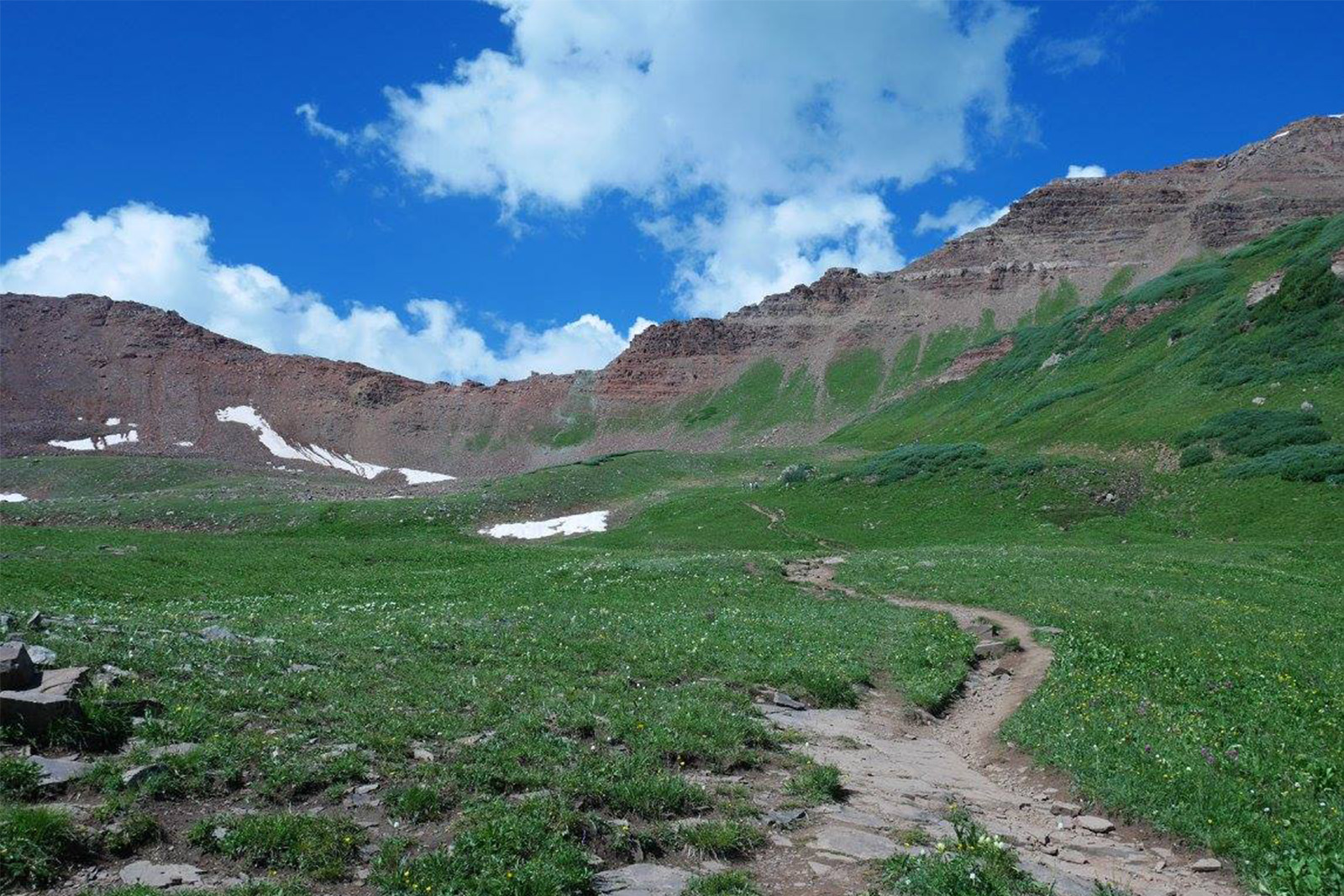 View of a ridge line from a trail. The area is open with no trees, the rocks are red in color, and the sky is a deep blue with some clouds.