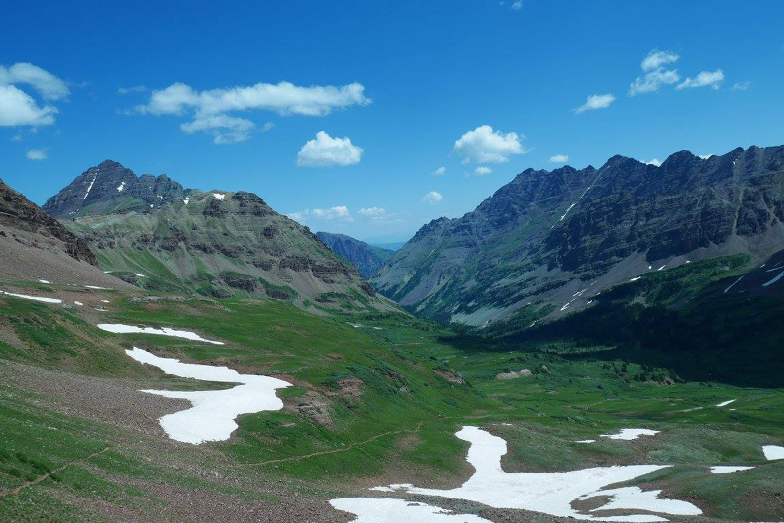 View of some ragged mountains with a trail running down into the valley. It is mostly open due to being above tree line with some patches of snow.